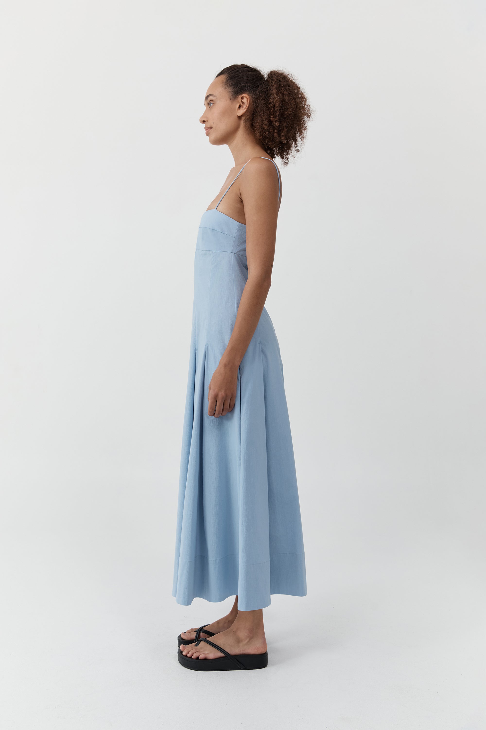 Side profile of a woman wearing the st. agni everyday cotton pleated dress in sky in front of a cream background