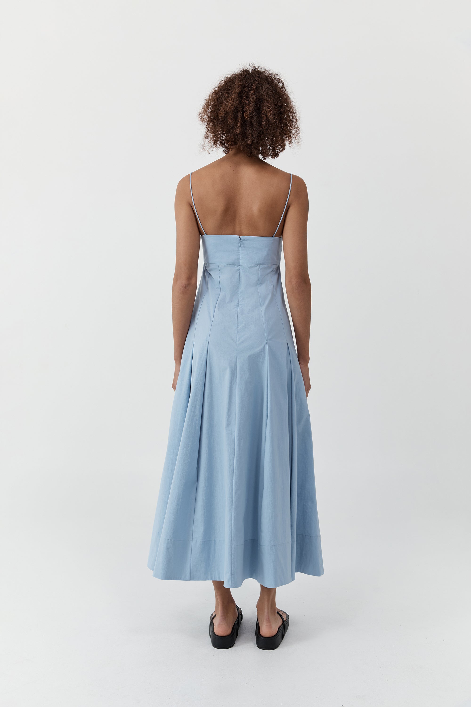 Backside of a woman wearing the st. agni everyday cotton pleated dress in sky in front of a cream background