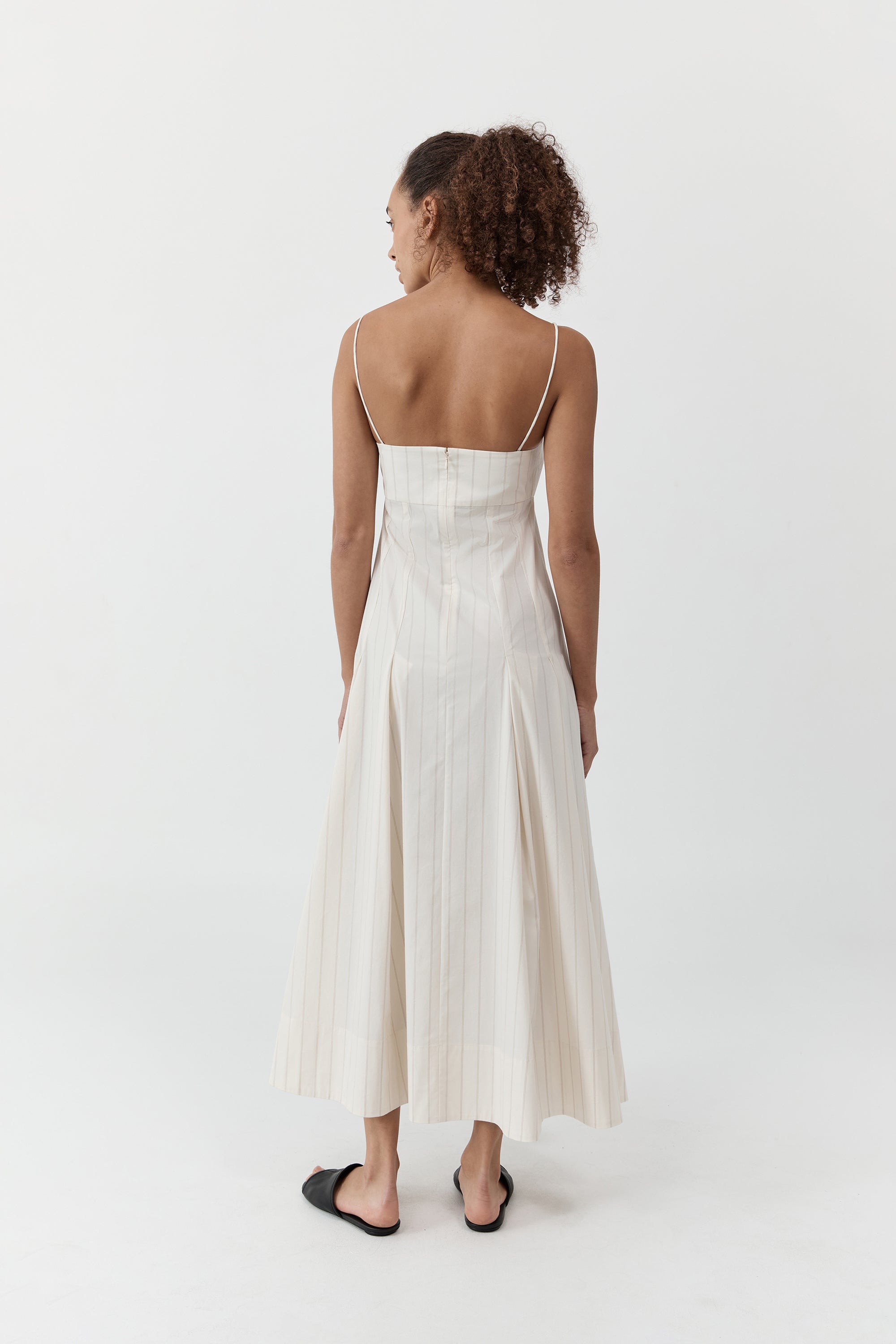 Backside of a woman wearing the st. agni everyday cotton pleated dress in ecru stripe standing in front of a cream background
