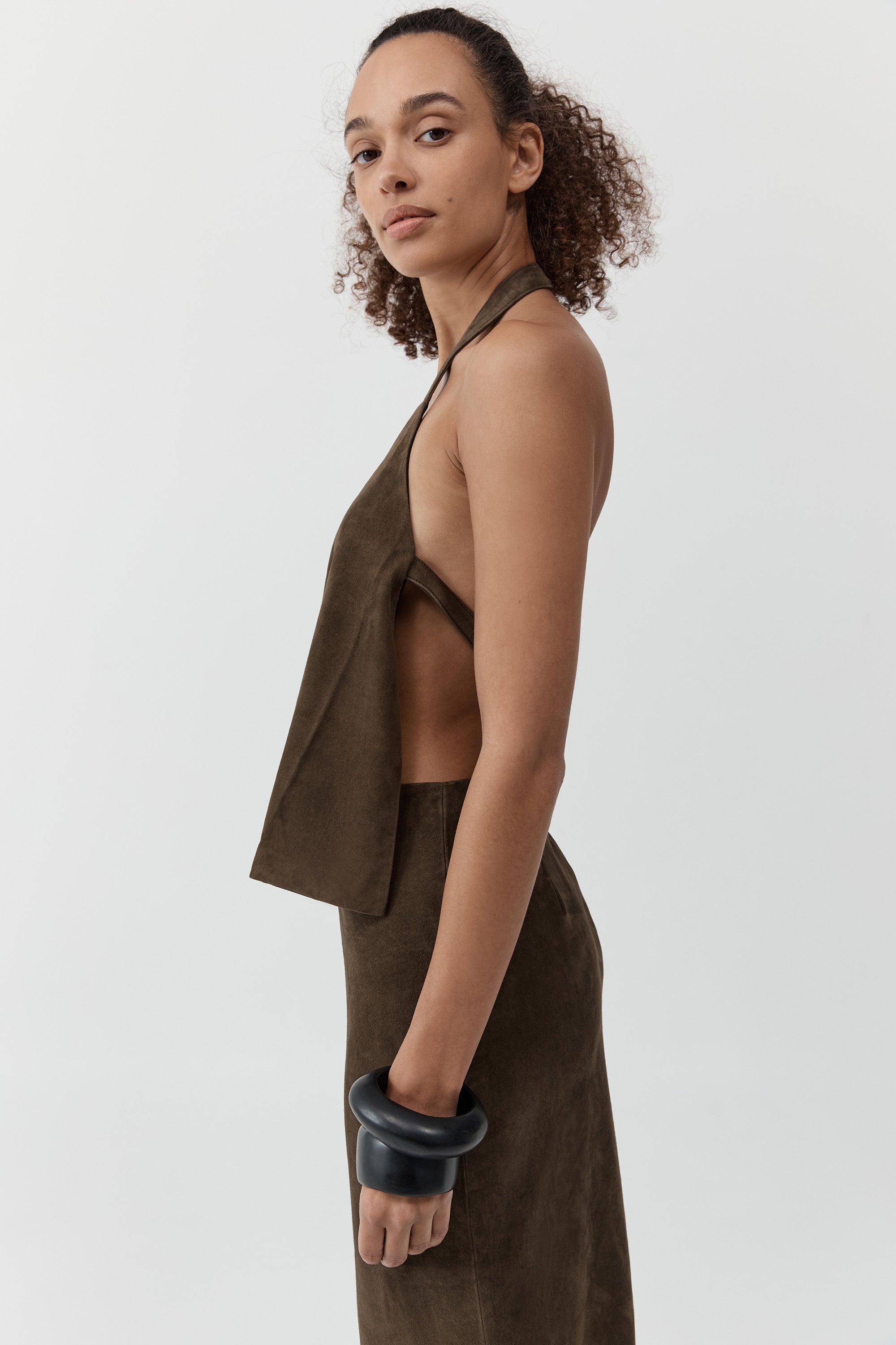 Side profile of a woman wearing a st. agni suede apron top with suede skirt in front of a cream background
