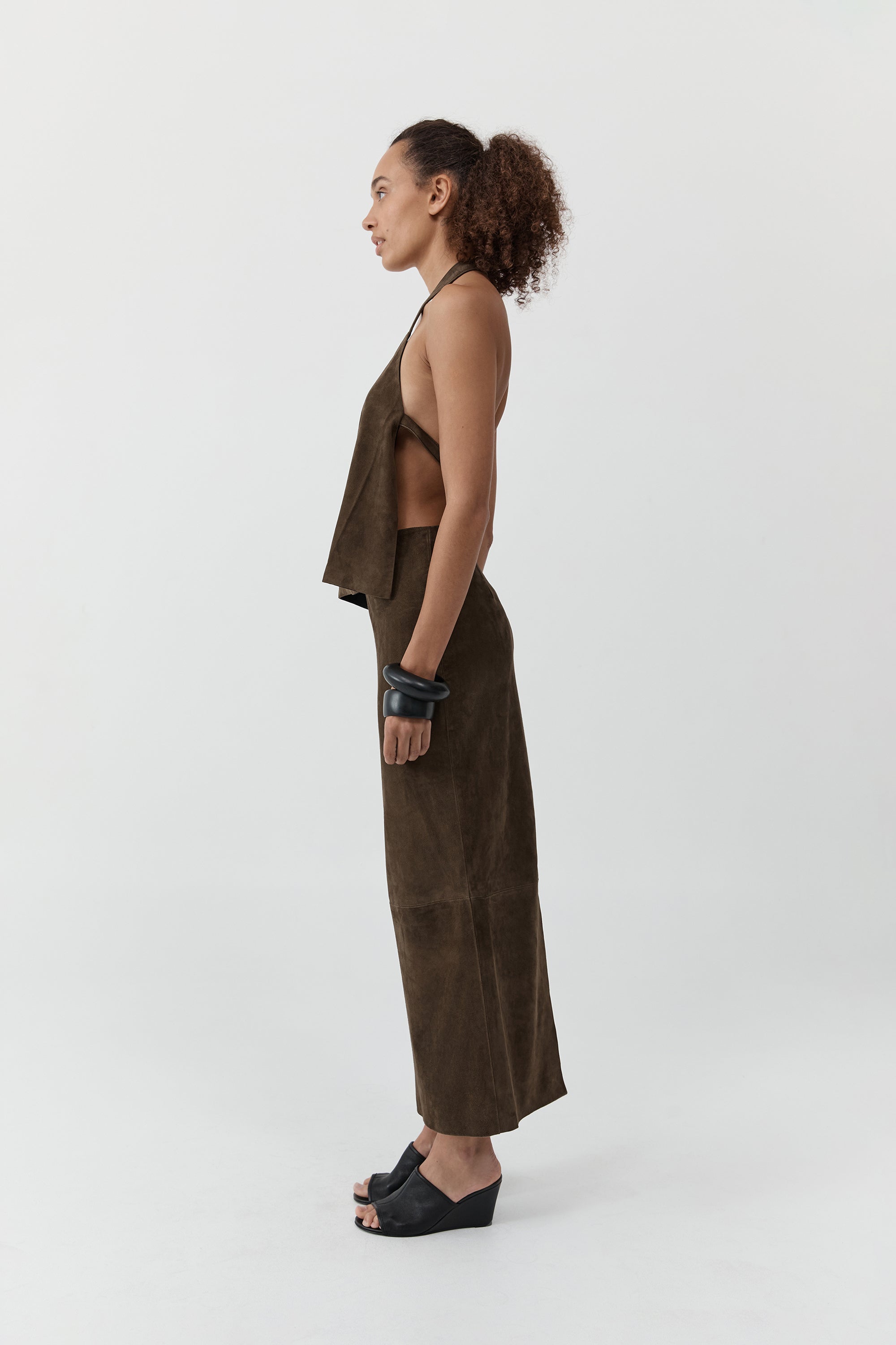 Side profile of a woman wearing a st. agni suede apron top with suede skirt in front of a cream background