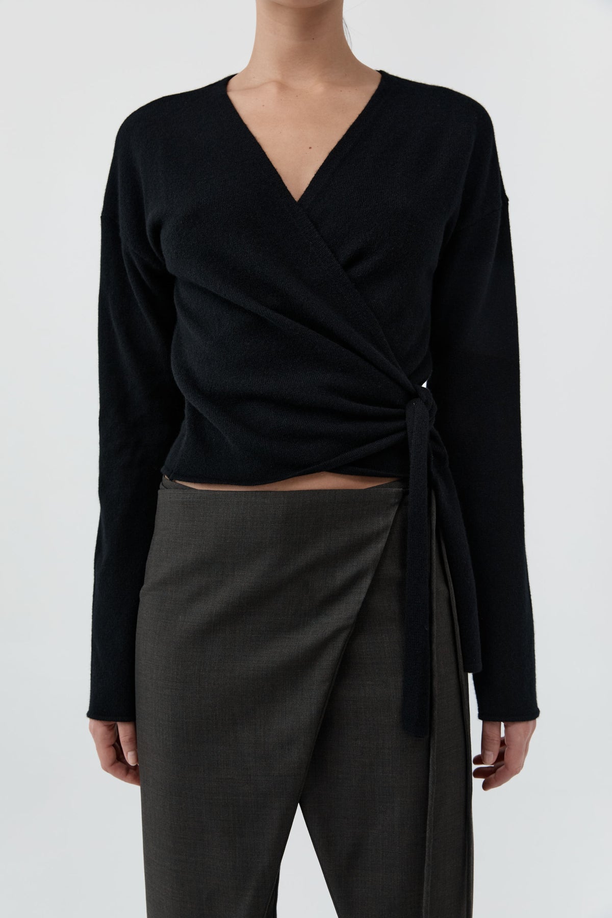Close up of a woman wearing St Agni Baby Wool Wrap Top and gray pants standing against a white background