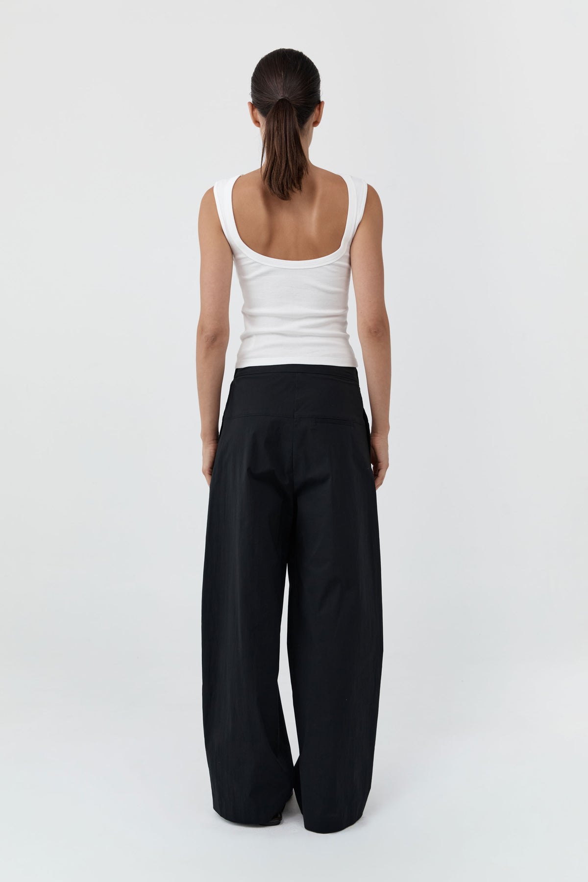 Backside of a woman wearing the St Agni Organic Cotton Ballerina Top-White and gray pants standing against a white background