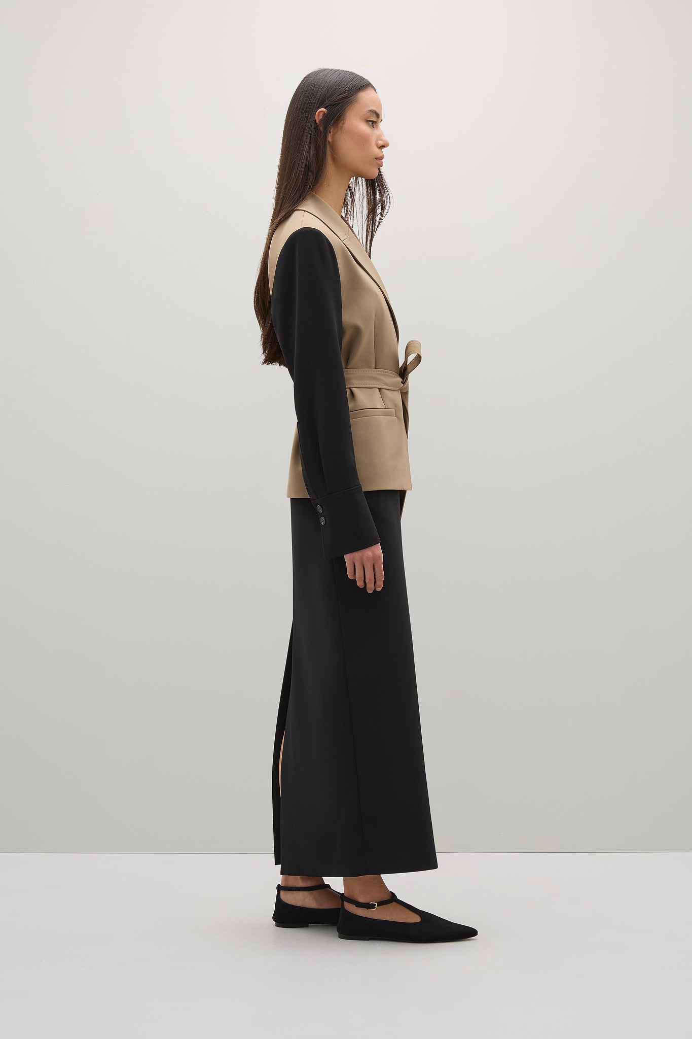 Woman wearing a beige vest over a black long-sleeve top and black pants on a white background