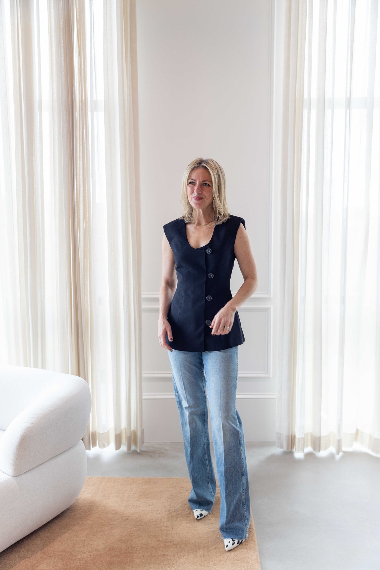 Woman wearing the house of dagmar sleeveless blazer and the amo marcenia mid-rise slouchy bootcut jean standing in a room with a white sofa and white curtains.