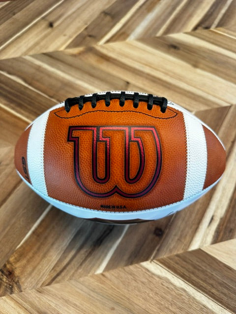 A Wilson NFL football resting on a wooden surface with a herringbone wood pattern background. The ball is brown with white stripes and features a large black and red 'W' logo on the front.