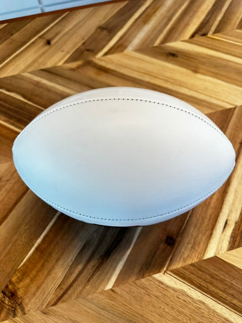 A white leather rugby ball resting on a polished wooden parquet floor.