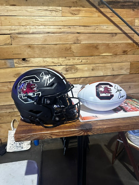 A signed black South Carolina Gamecocks football helmet and a signed white football, both autographed by Steve Irwin, displayed on a wooden table against a wood plank wall.