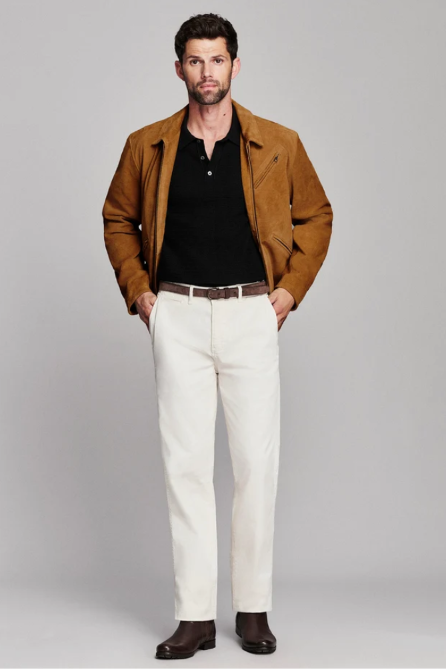 A man modeling a brown suede jacket over a black henley shirt, paired with off-white trousers, a brown belt, and dark brown shoes, shot against a solid gray studio background.