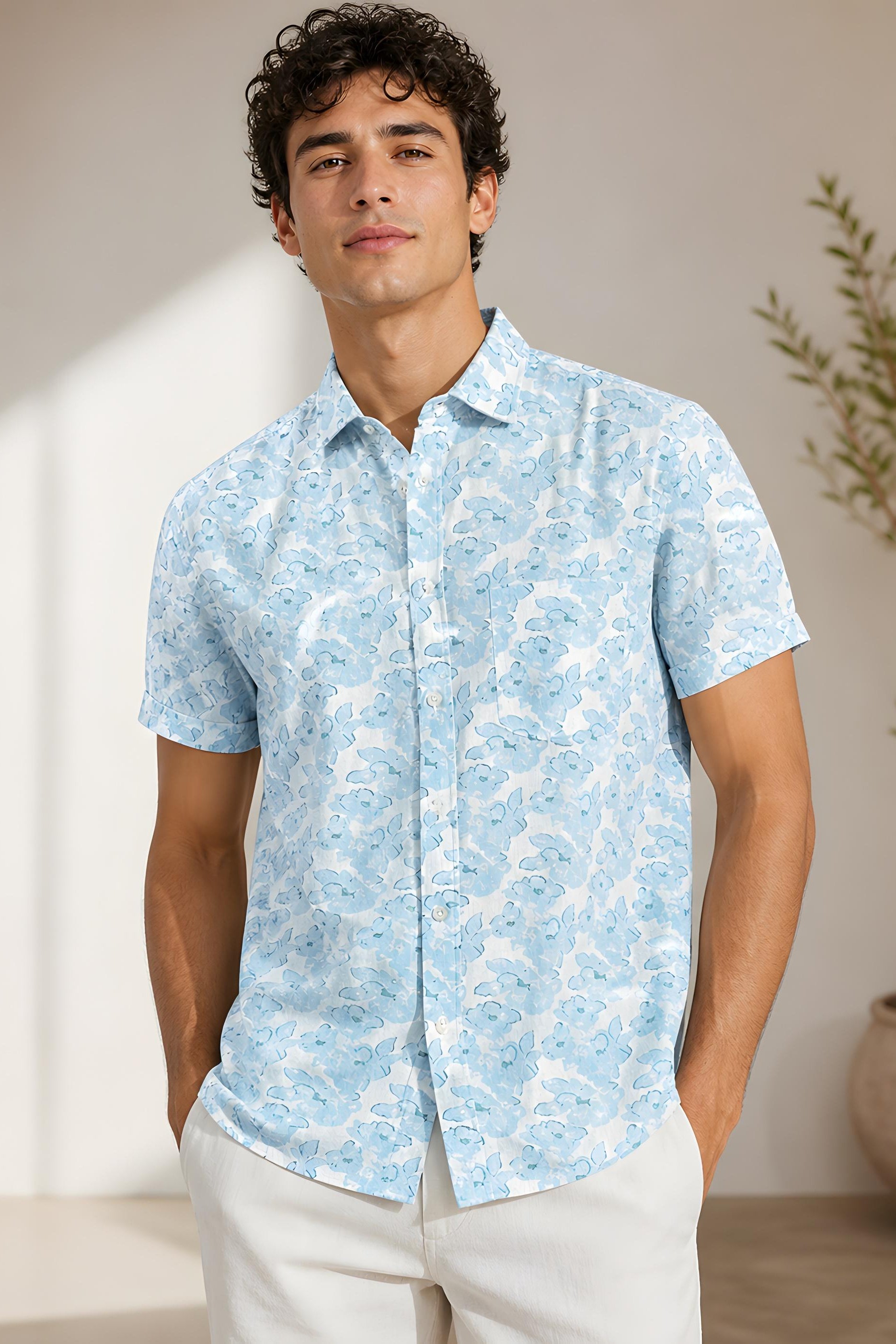 Man wearing a light blue patterned shirt standing in a room with a plant.