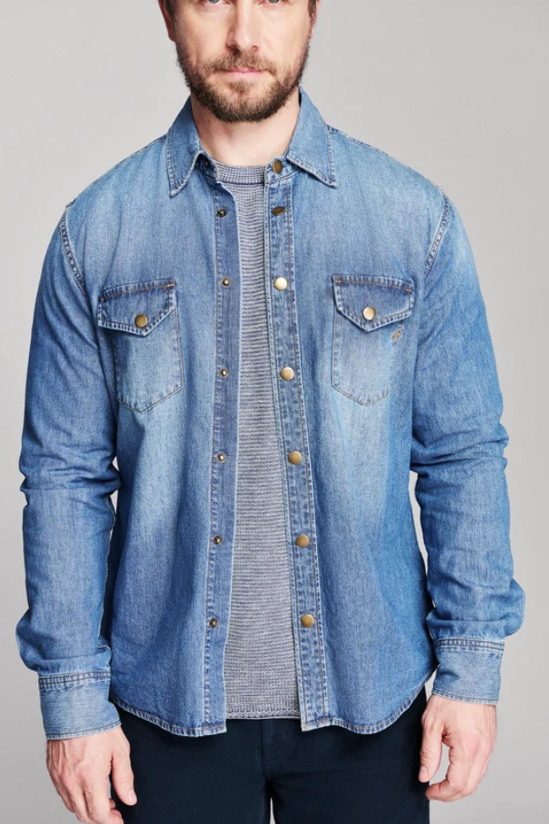 A man wearing a light blue denim button-up shirt jacket over a gray striped t-shirt and dark pants, photographed against a solid gray studio background.