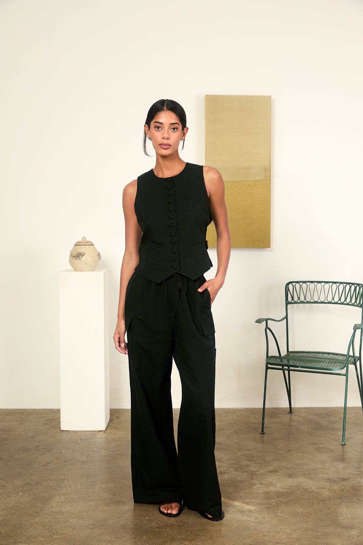 Woman in a black outfit standing in a minimalistic room with a chair and decorative items.