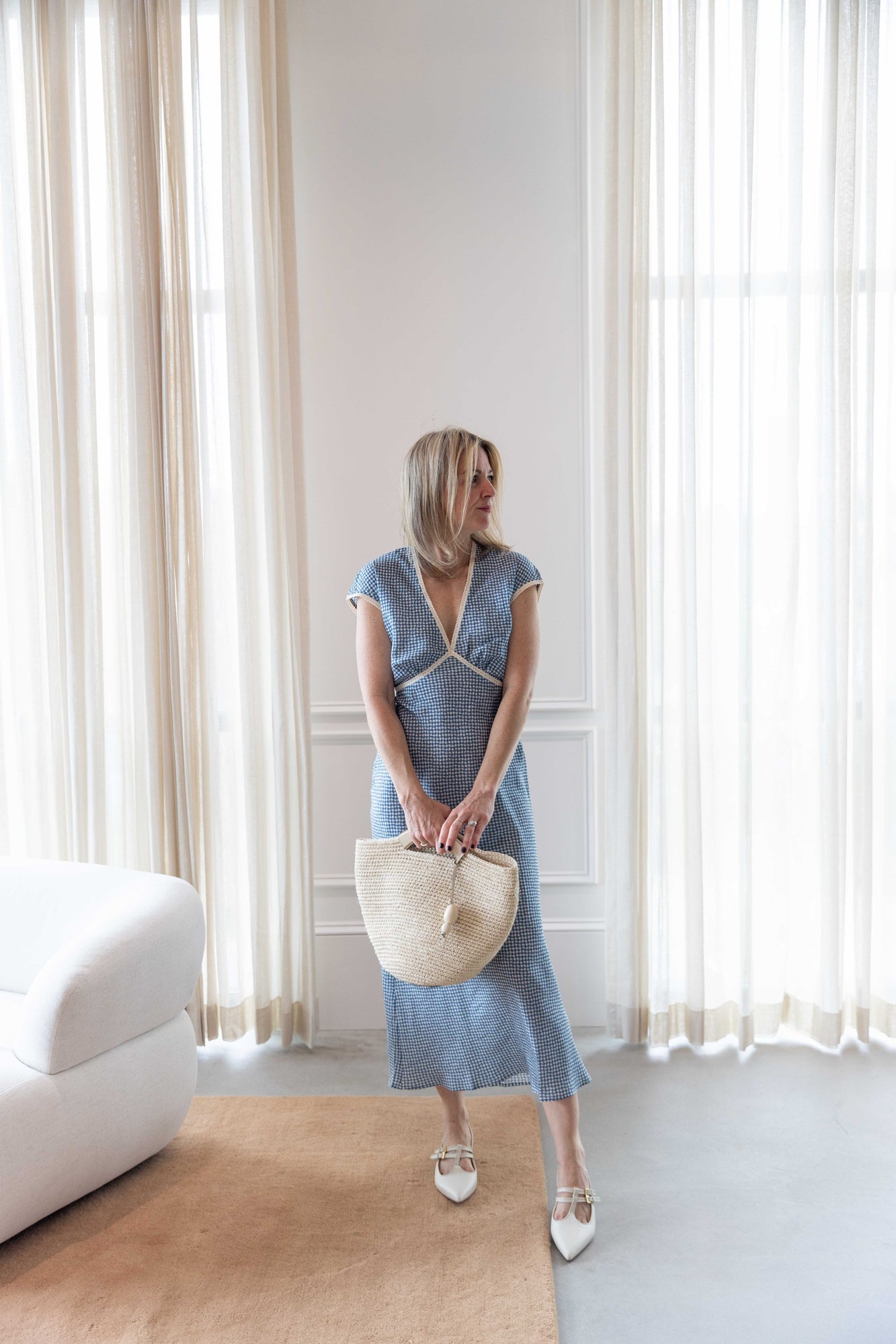 Woman in the ciao lucia indine dress holding the hunting season large woven shopper and standing in a room with white curtains.