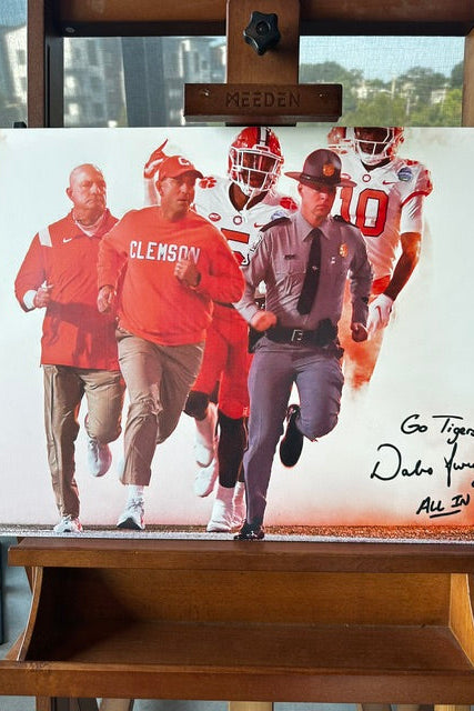 A signed canvas print of Clemson coacj dabo swinney running alongside players and a police officer, displayed on a wooden easel. The print features a white background with a printed collage of the subjects in red and gray, with a black autograph in the bottom right corner reading 'Go Tigers! Dabo Swinney All In'.