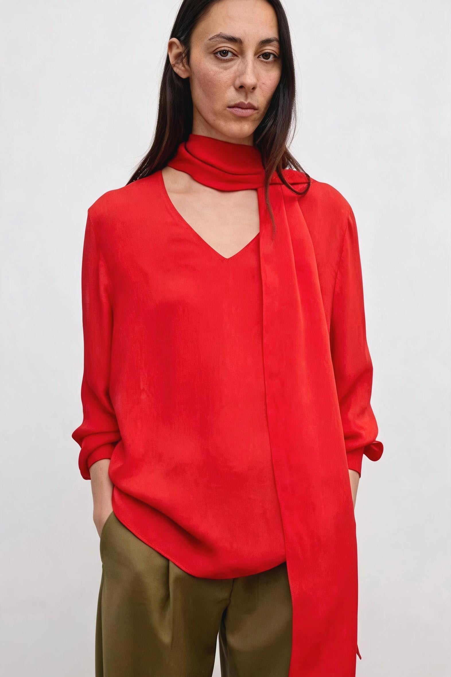 A woman wearing a vibrant red long-sleeve top with a high neck and a draped tie detail, paired with olive green trousers, against a plain light gray background.