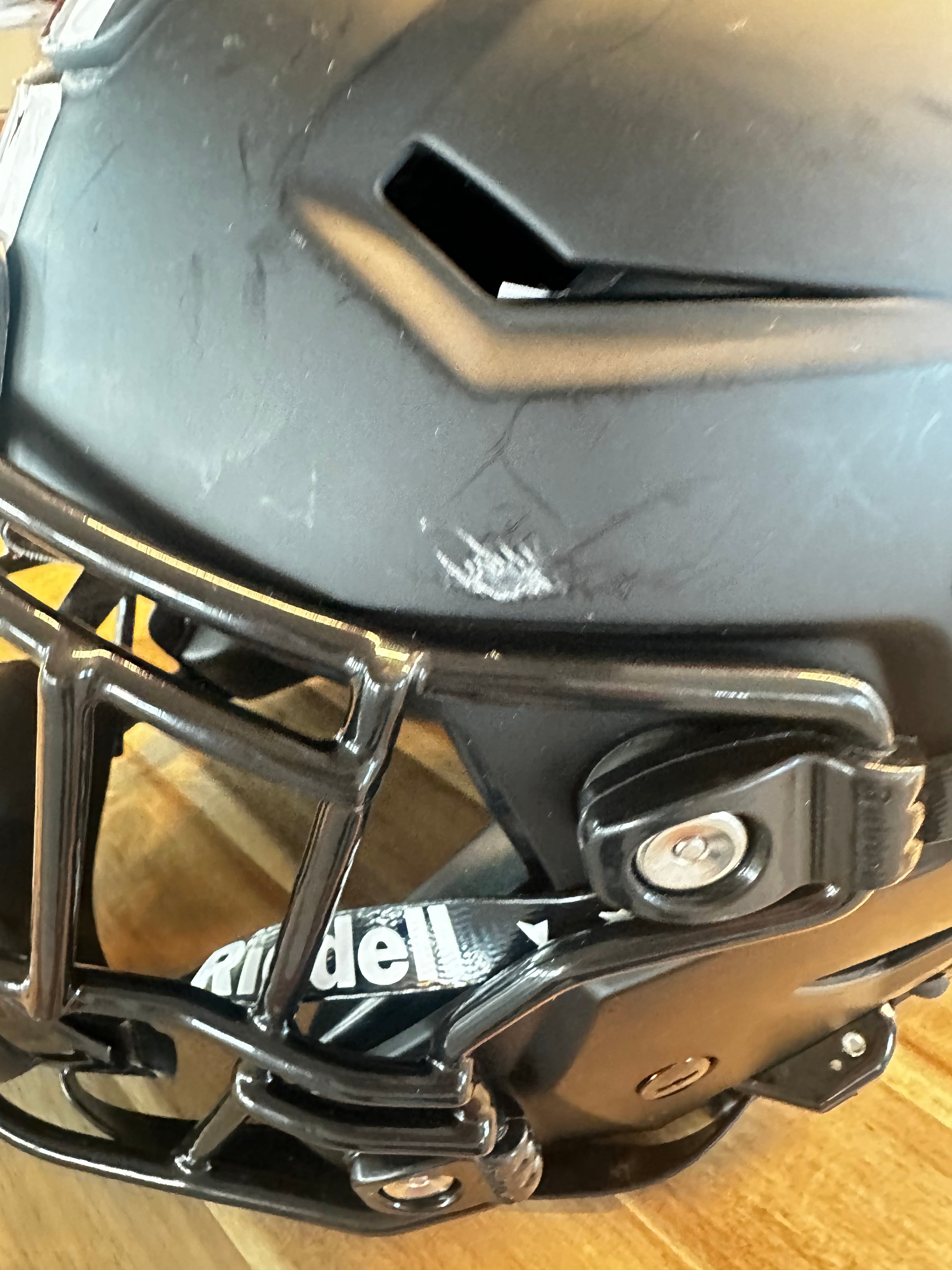 Close-up of a black Riddell football helmet with a faceguard, resting on a wooden floor. The helmet shows visible scuffs and scratches on the shell.