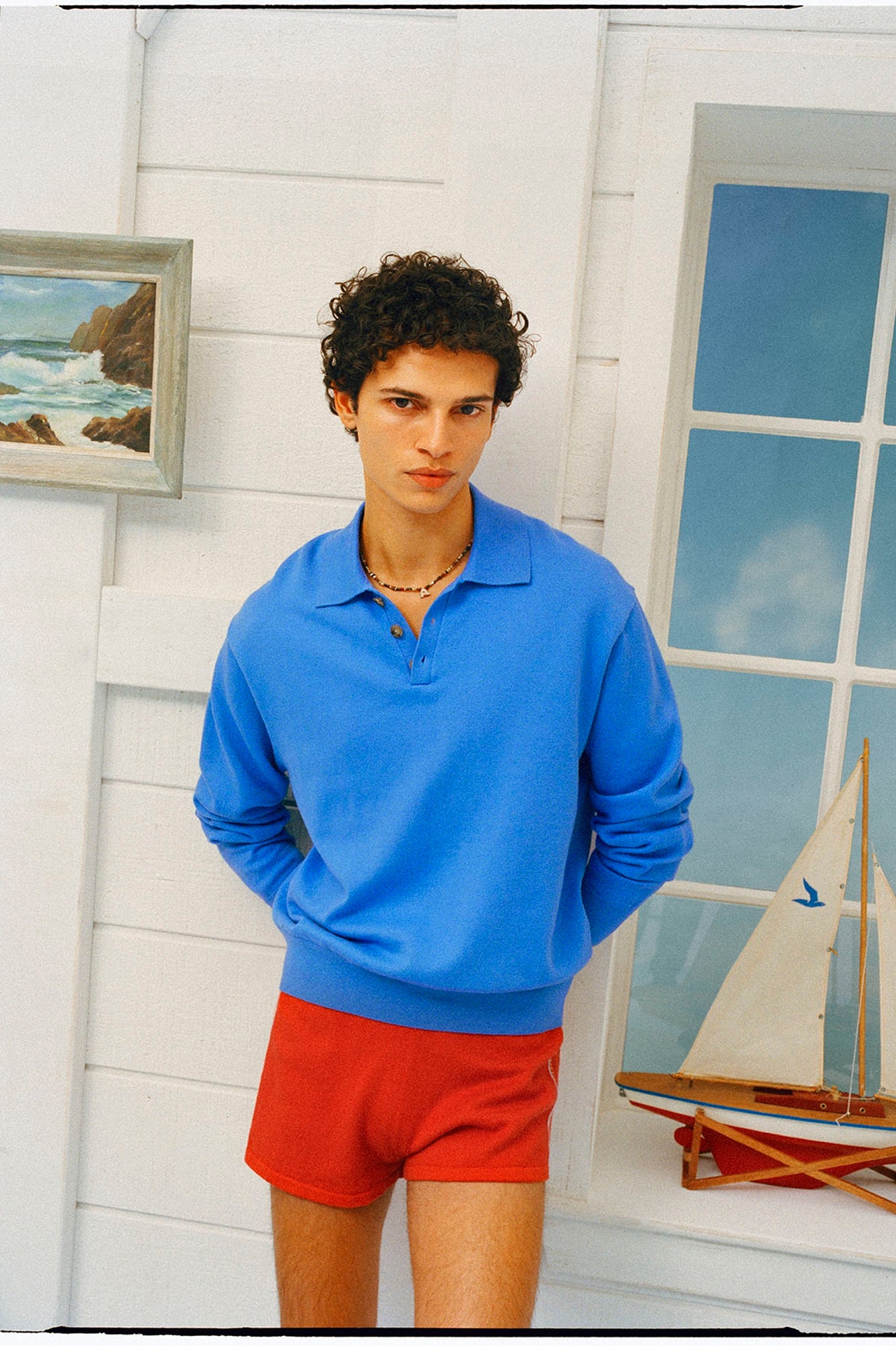 Person wearing a blue sweater and red shorts standing in a room with a sailboat model and framed picture on the wall.