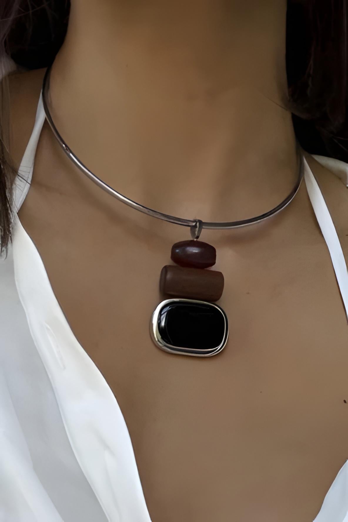 A close-up of a person's neck and chest wearing a minimalist silver wire choker necklace with stacked wooden beads and a large black rectangular stone pendant.