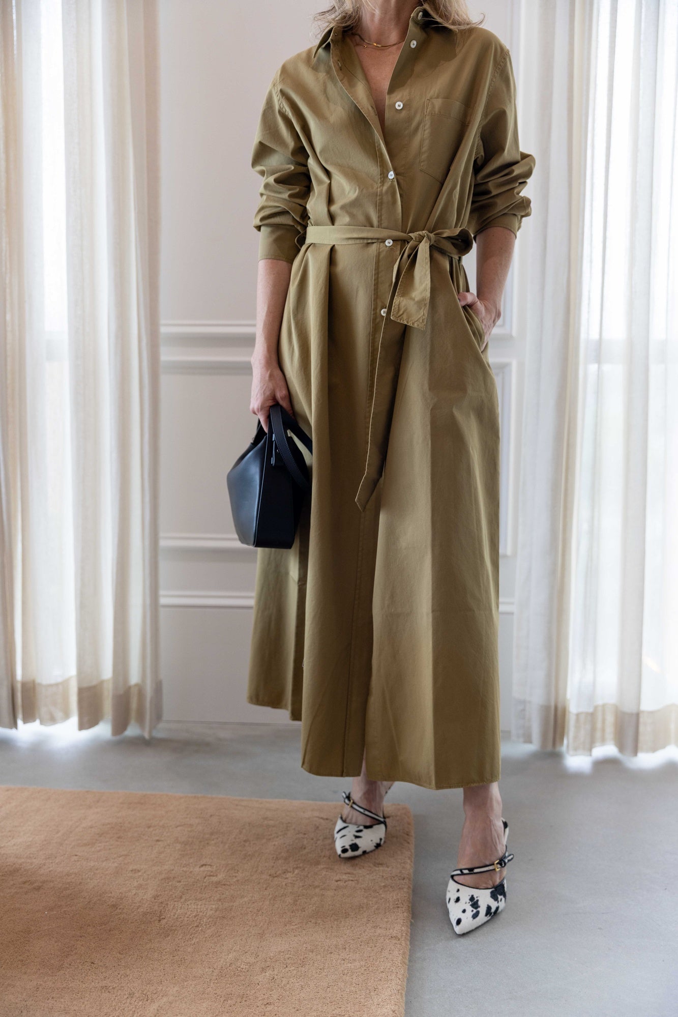 Close up of a woman wearing the green house of dagmar belted shirt dress standing in a room with a tan area rug and white curtains.