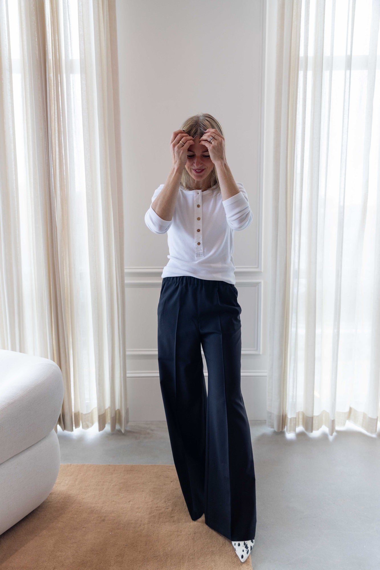 Woman wearing a house of dagmar henley top in white and house of dagmar elastic trouser standing in a room with white curtains.