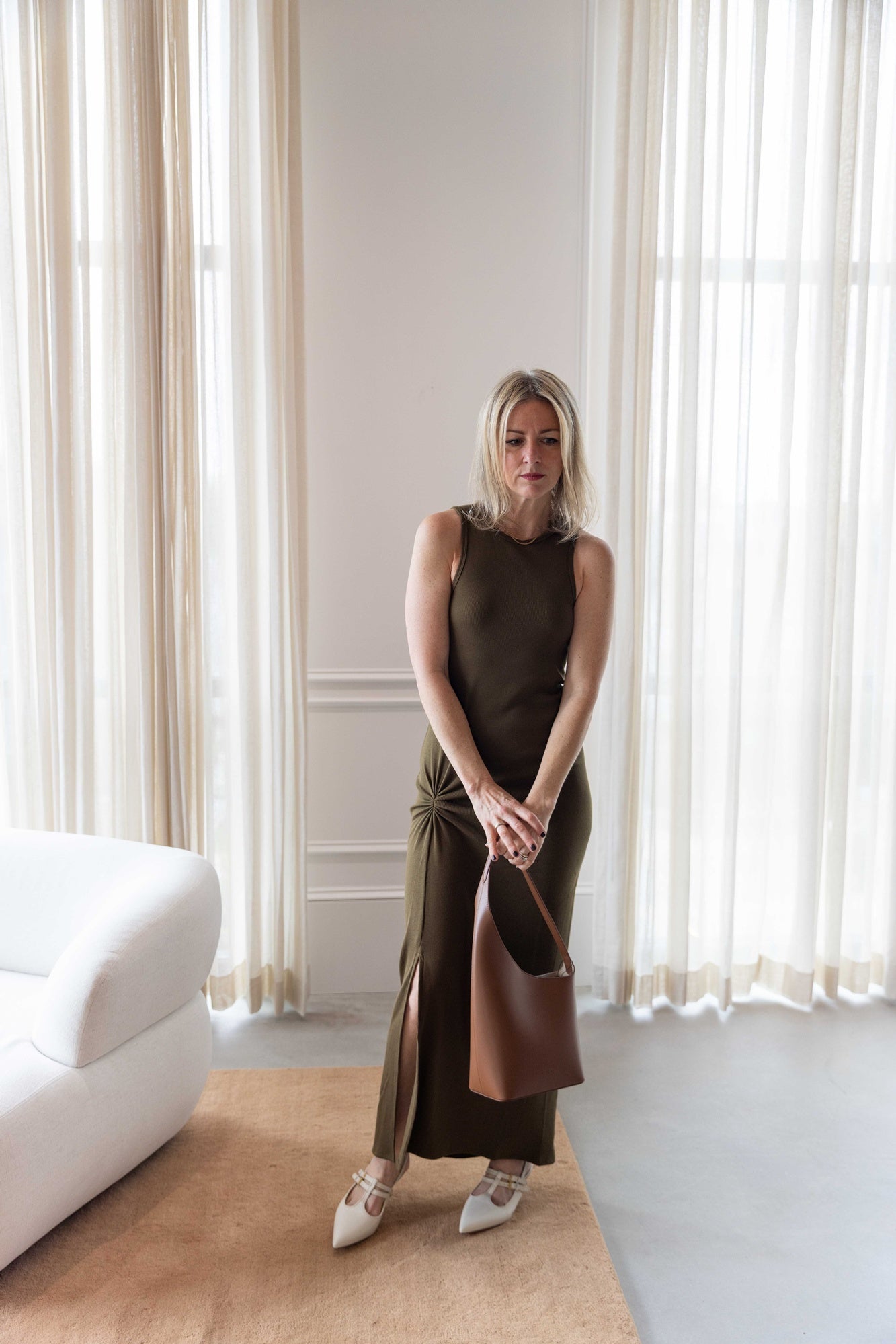 Woman wearing the house of dagmar knot tank dress and holding the aesther ekme new demi lune handbag house standing in a room with white curtains and a white sofa.