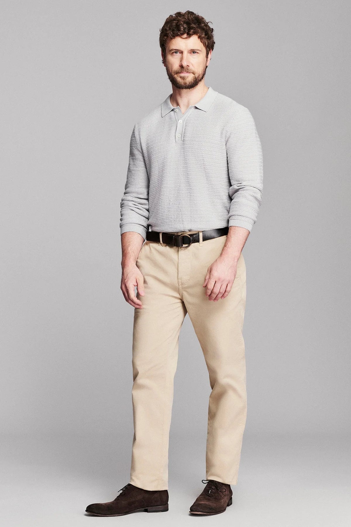 A man modeling a light gray long-sleeve polo shirt, beige trousers, a black belt, and dark brown dress shoes against a solid gray studio background.