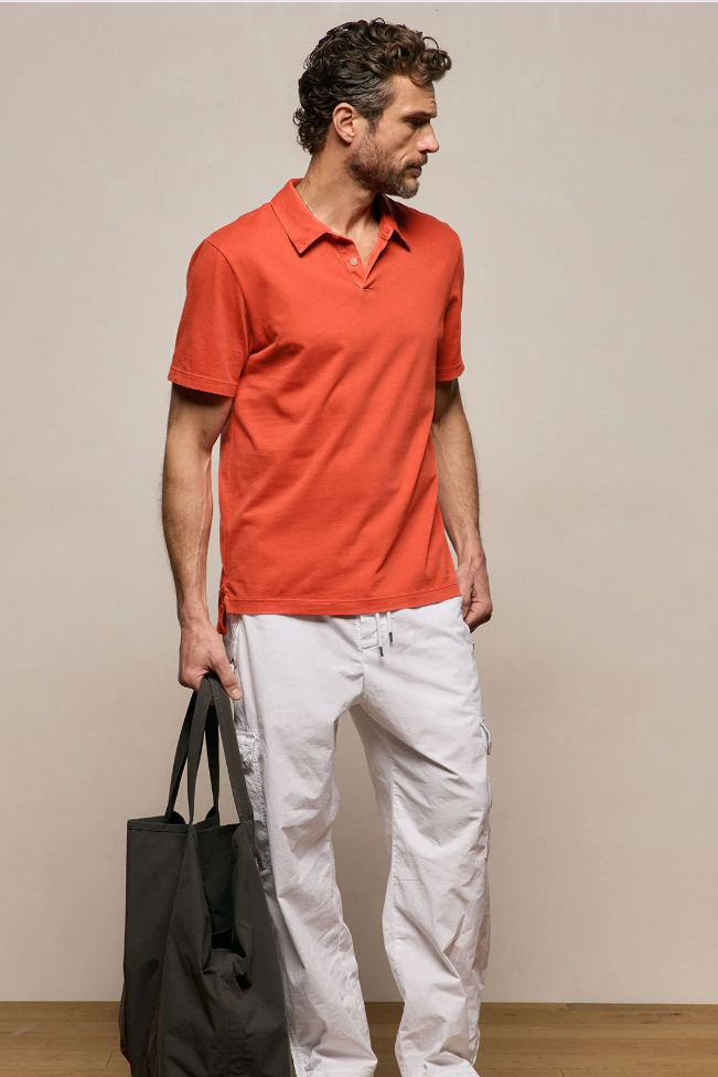 Man wearing an orange polo shirt and white pants holding a black bag against a beige background