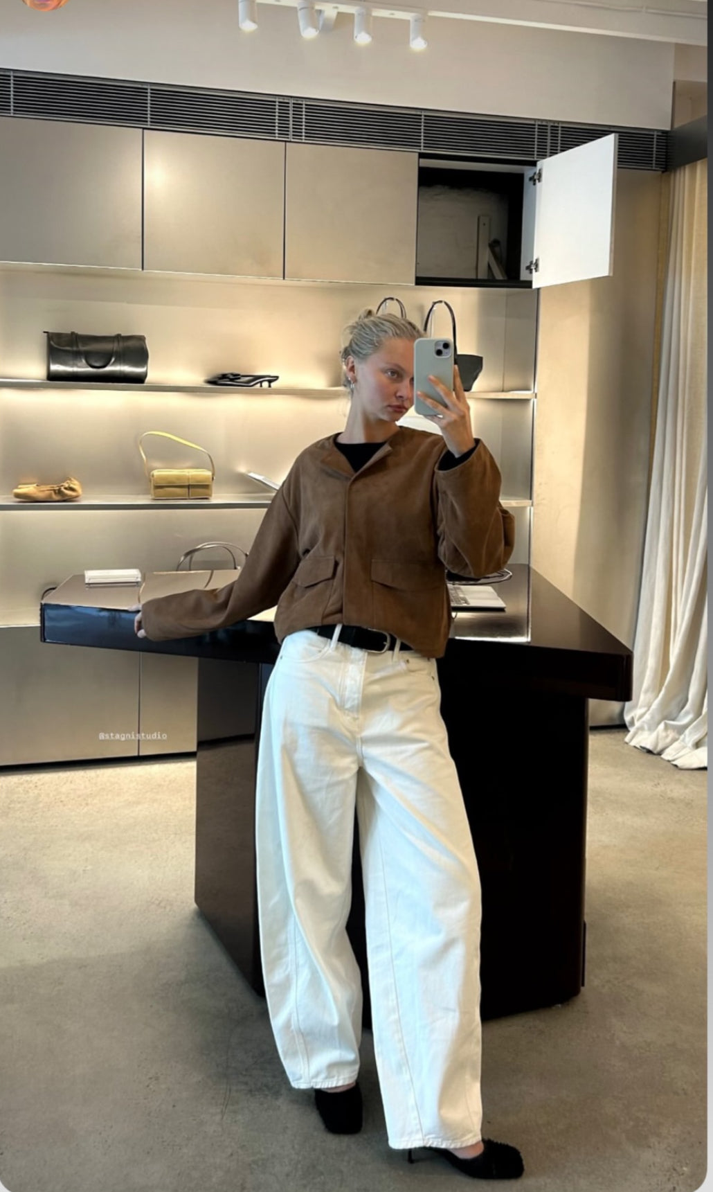 Person taking a mirror selfie in a modern interior setting wearing a brown jacket and white pants.