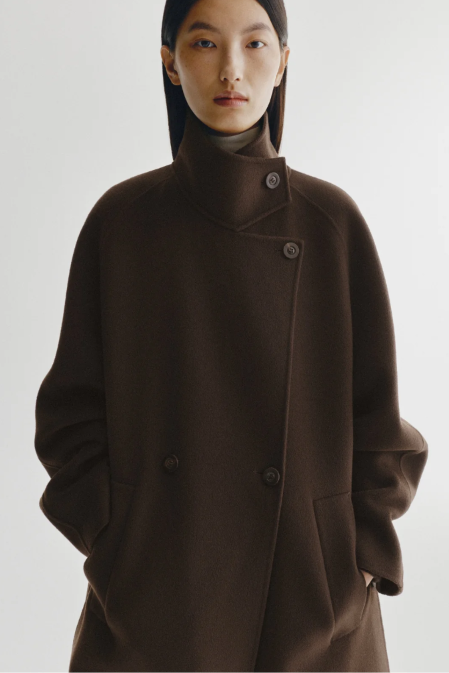 A model wearing a dark brown wool-blend coat with a high mock neck and asymmetrical button closure, styled with a matching brown head covering against a light gray studio background.