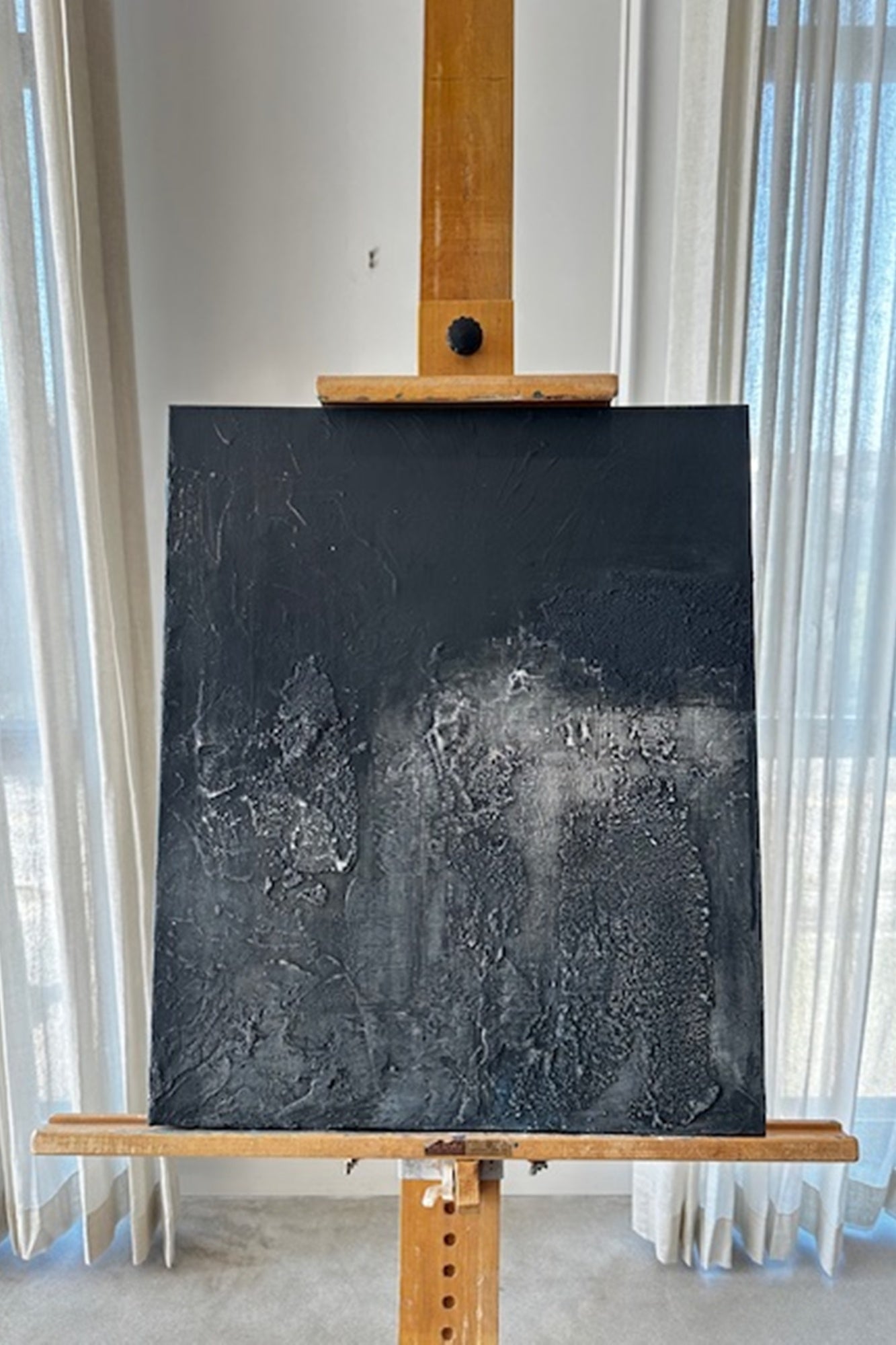Black textured painting on a wooden easel against a white curtain background