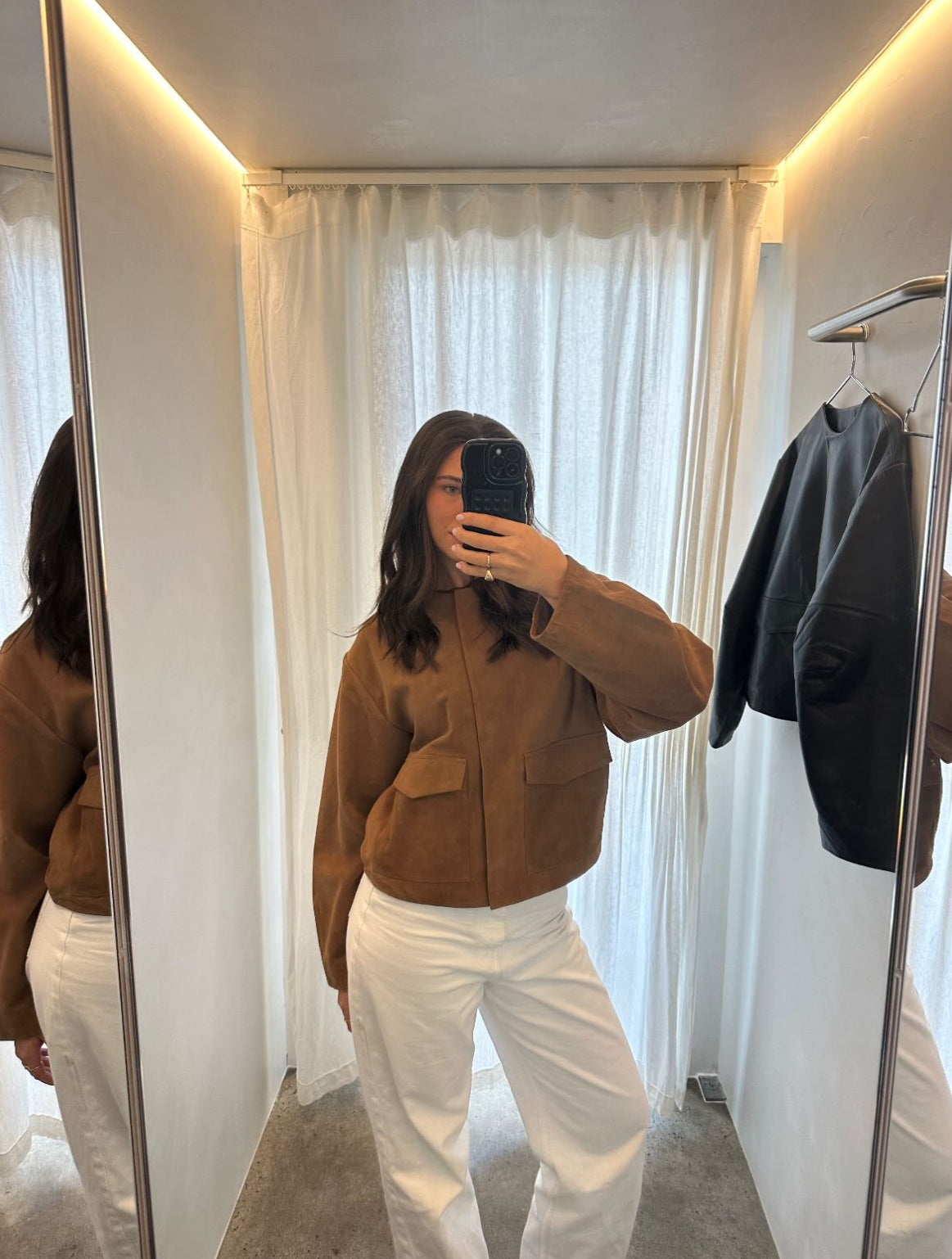 Person wearing a brown jacket and white pants taking a mirror selfie in a room with white curtains and a black jacket on a rack.