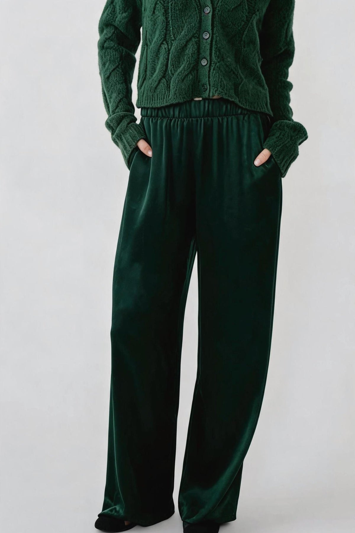 A person wearing a dark forest green knit cardigan and matching wide-leg velvet pants, standing against a light gray studio background with hands in the pants pockets.