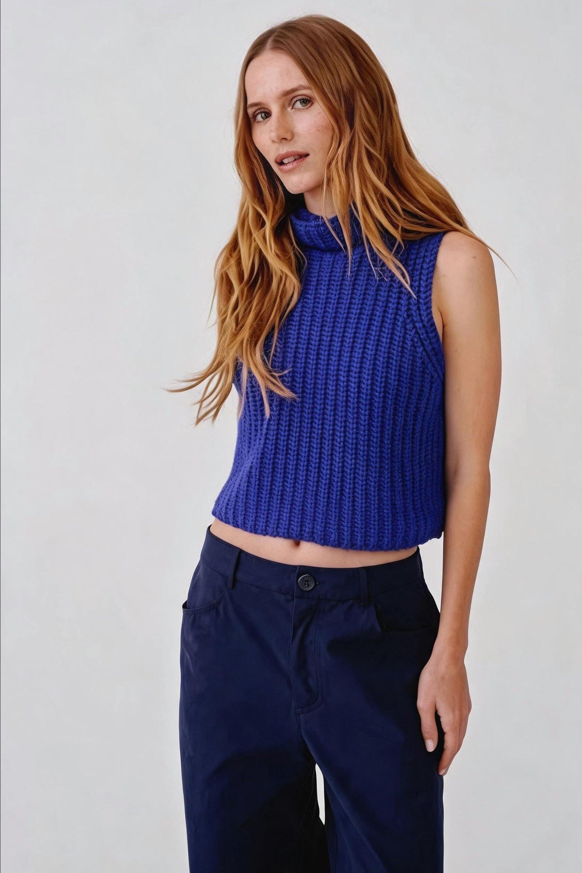 A woman with long light brown hair wearing a textured royal blue sleeveless mock-neck crop top and dark navy trousers, standing against a light gray studio background.