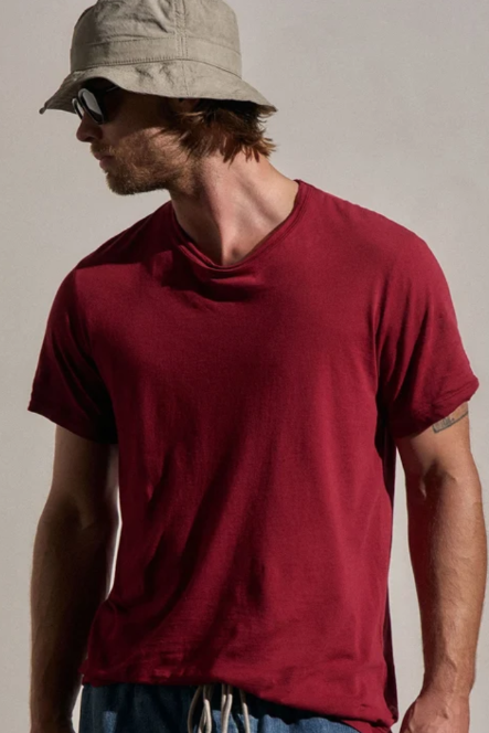 A man wearing a solid dark red v-neck t-shirt, a beige bucket hat, and sunglasses, photographed against a neutral light beige background.