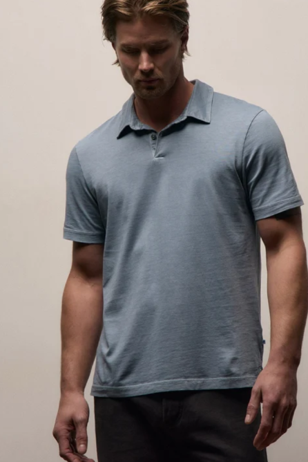 A man wearing a light slate blue short-sleeve polo shirt and dark pants, standing against a plain beige studio background.