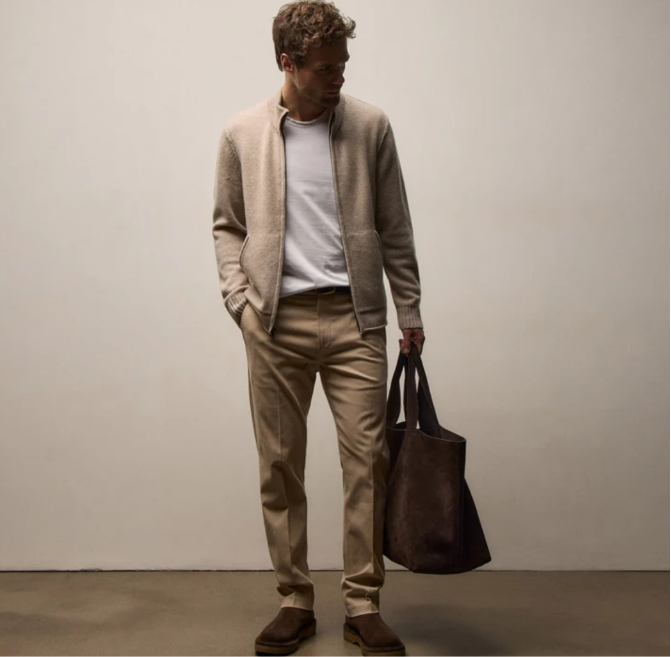 A man wearing a beige knit zip-up cardigan over a white t-shirt, khaki trousers, and brown suede boots, holding a dark brown leather tote bag against a plain light gray studio background.