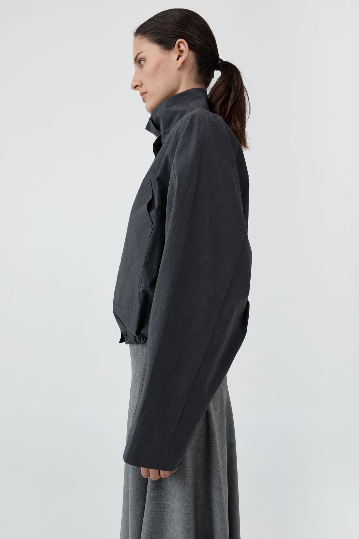 A woman shown in profile wearing a dark charcoal gray cropped jacket with a high collar and long sleeves, paired with a matching gray skirt, against a plain light gray studio background.