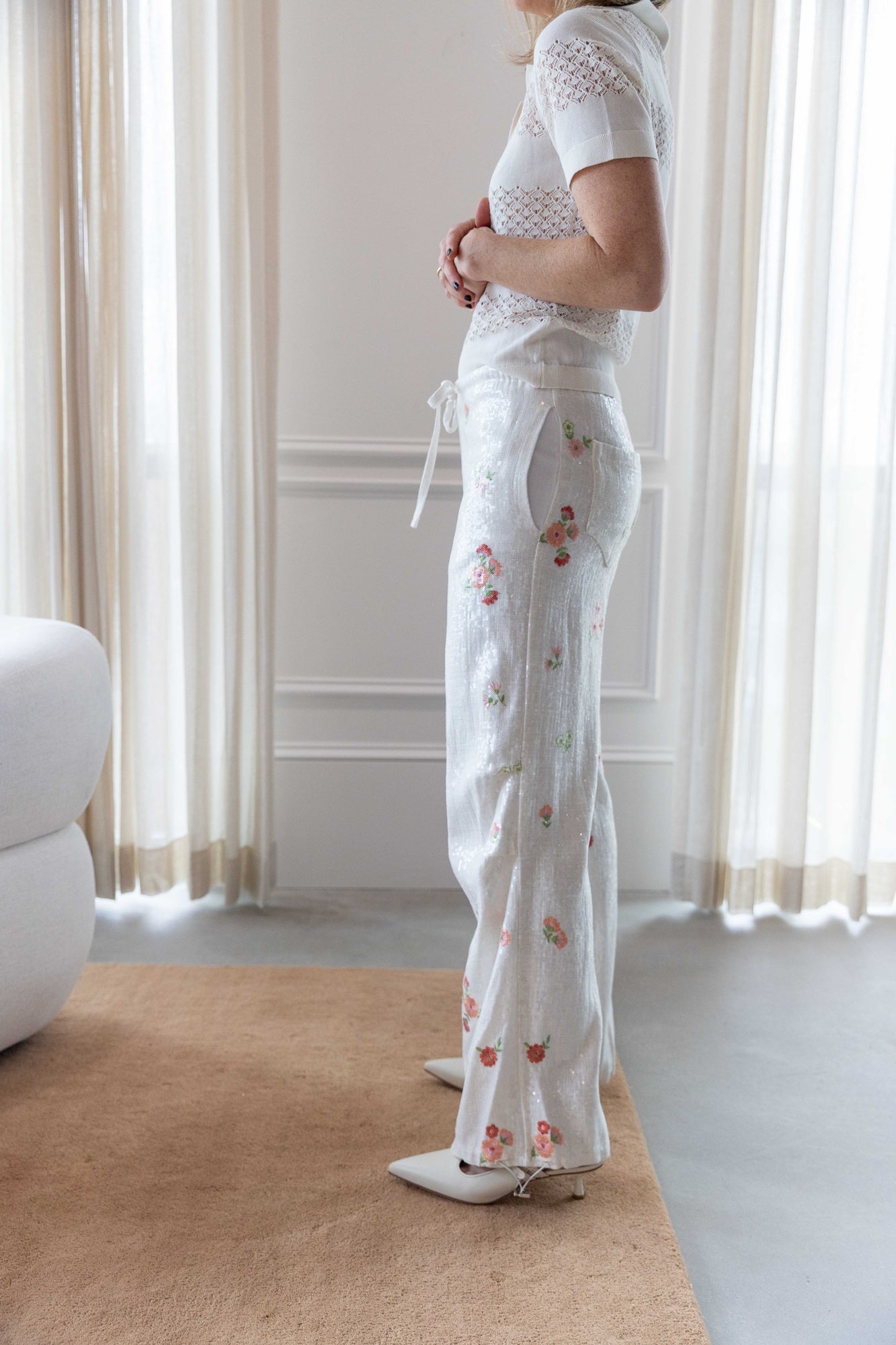 Woman wearing the garment tanzania logo polo and the alfie grace sequin pant standing in a room with white curtains.