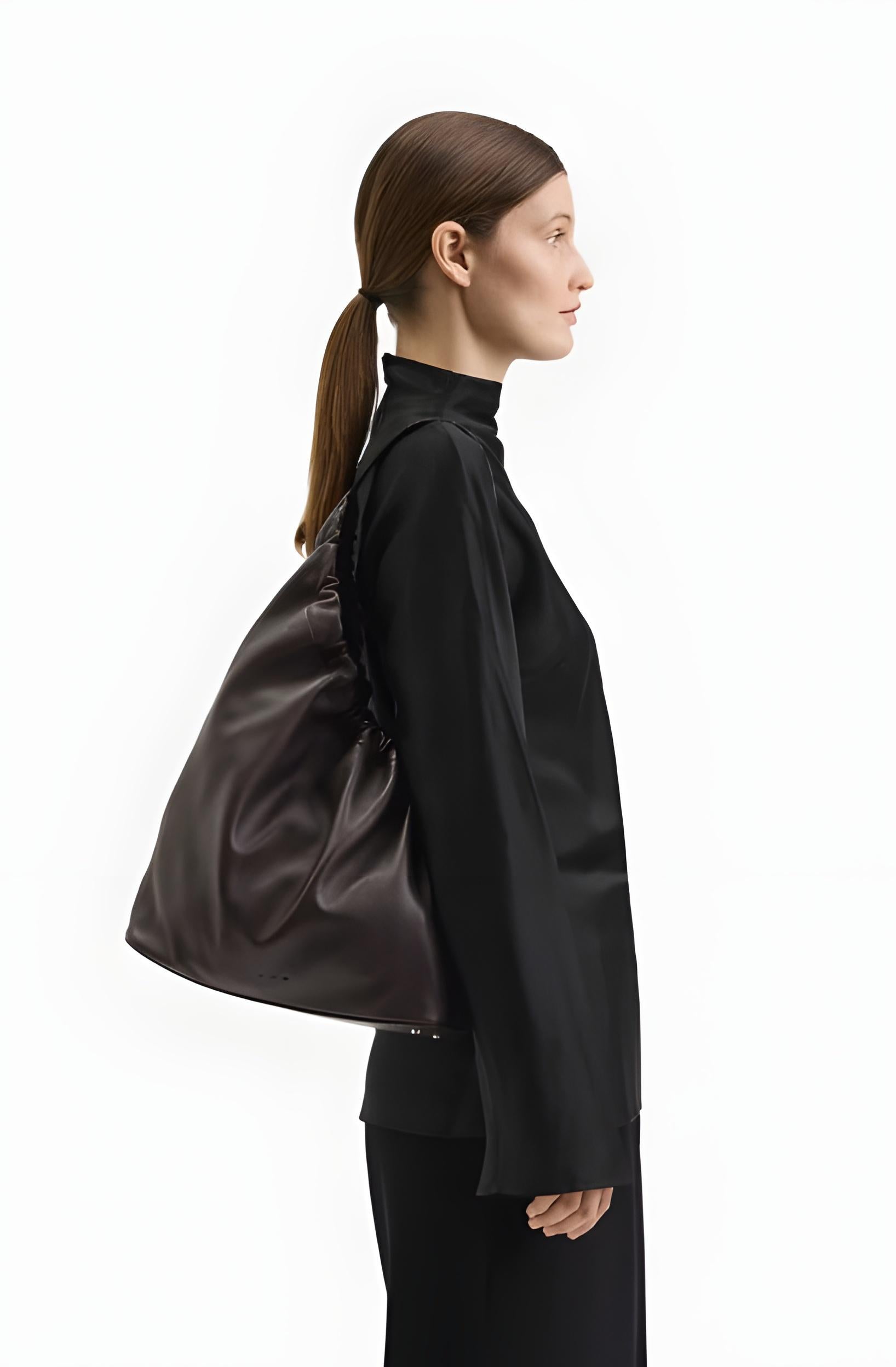 A woman in a black mock-neck top and black pants is shown in profile carrying a large, dark brown leather shoulder bag against a plain white background.