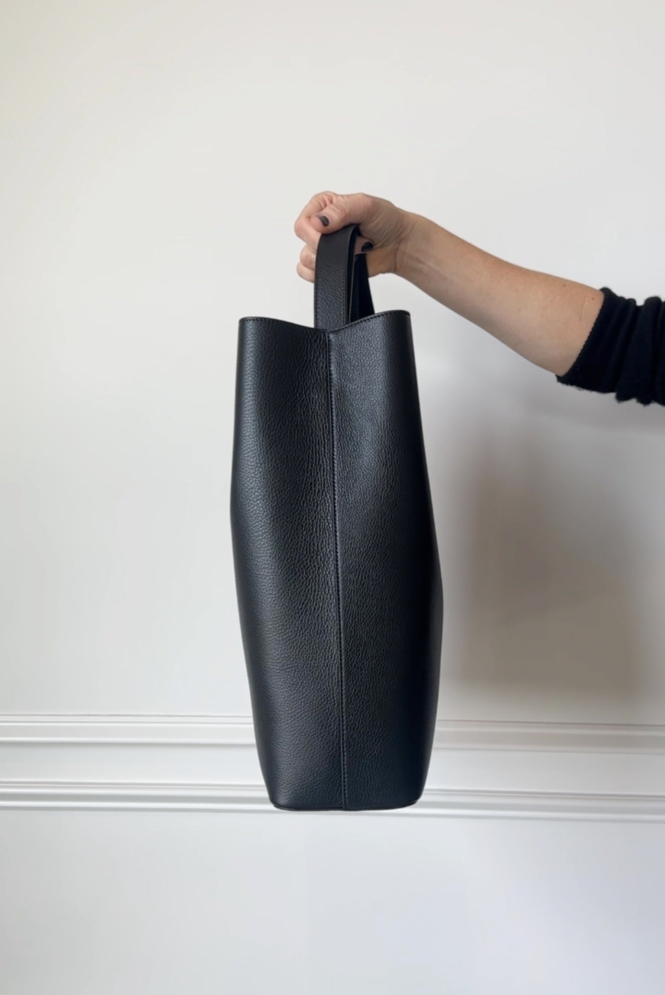 Black leather tote bag held by a hand against a white wall