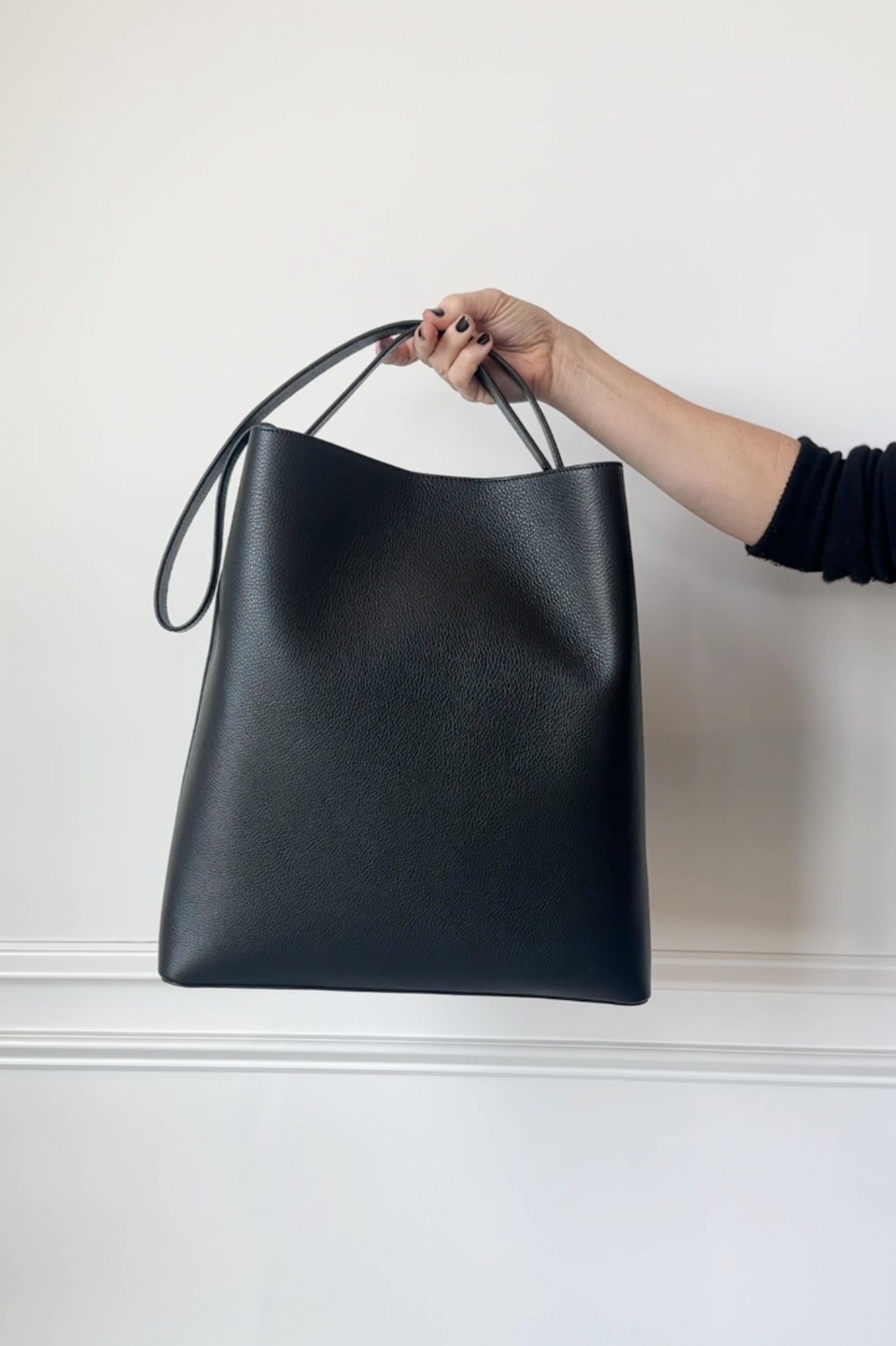 Black leather tote bag held by a hand against a white background