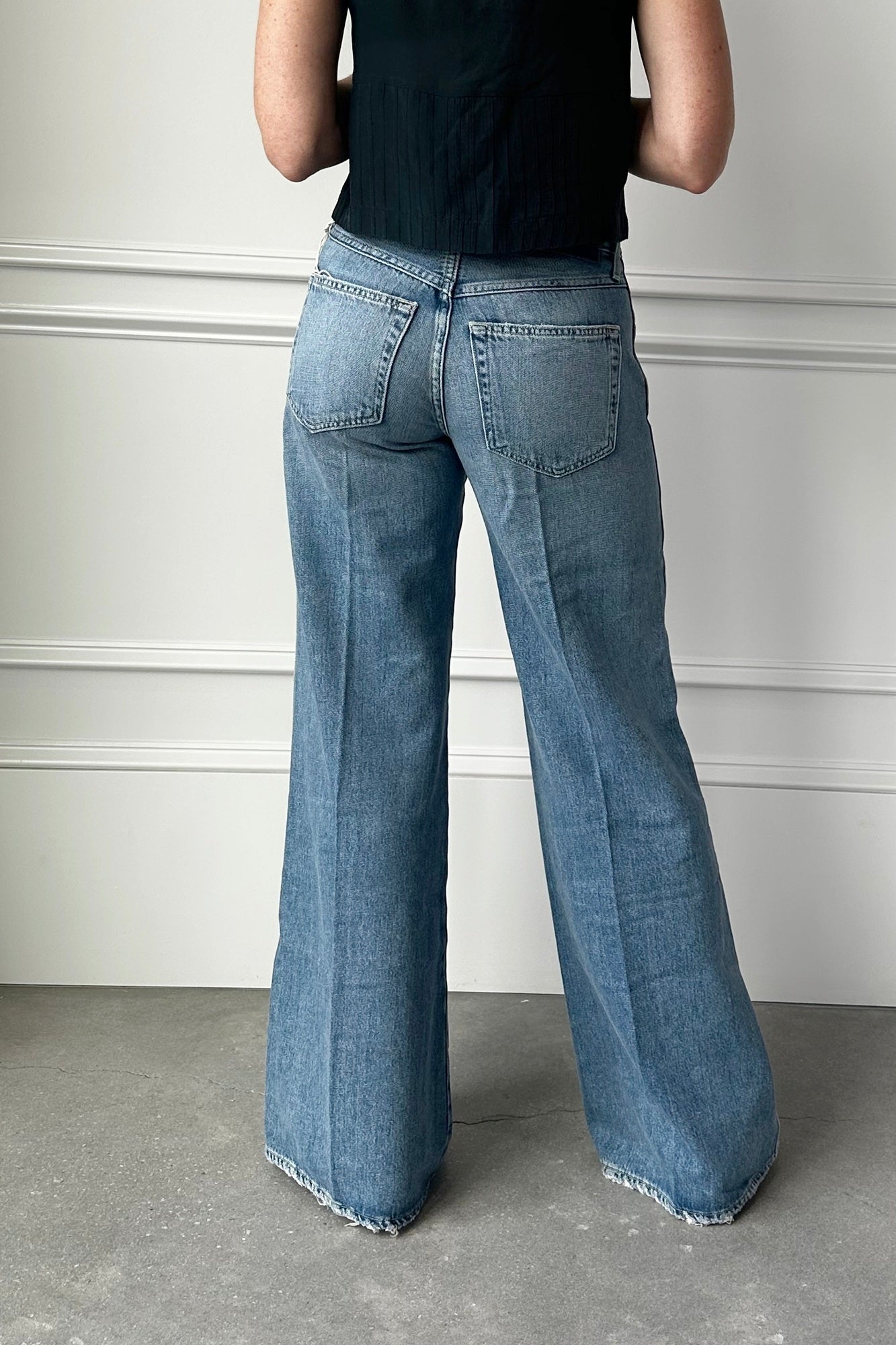 Person wearing blue flared jeans and a black top against a white wall.