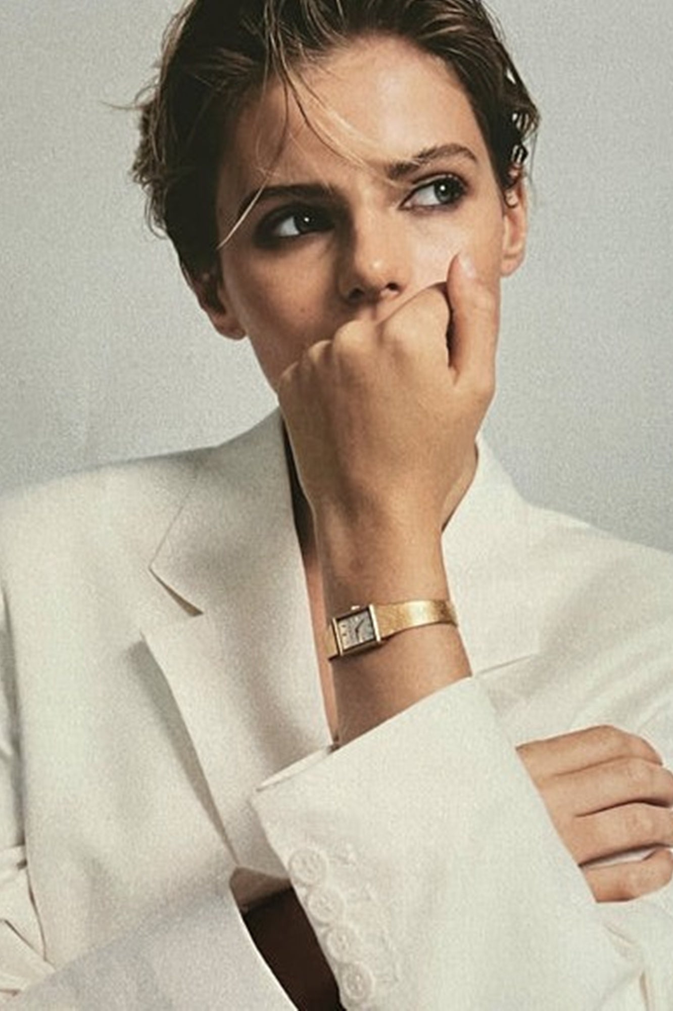 Woman in a white suit with a gold watch on her wrist, posing thoughtfully.