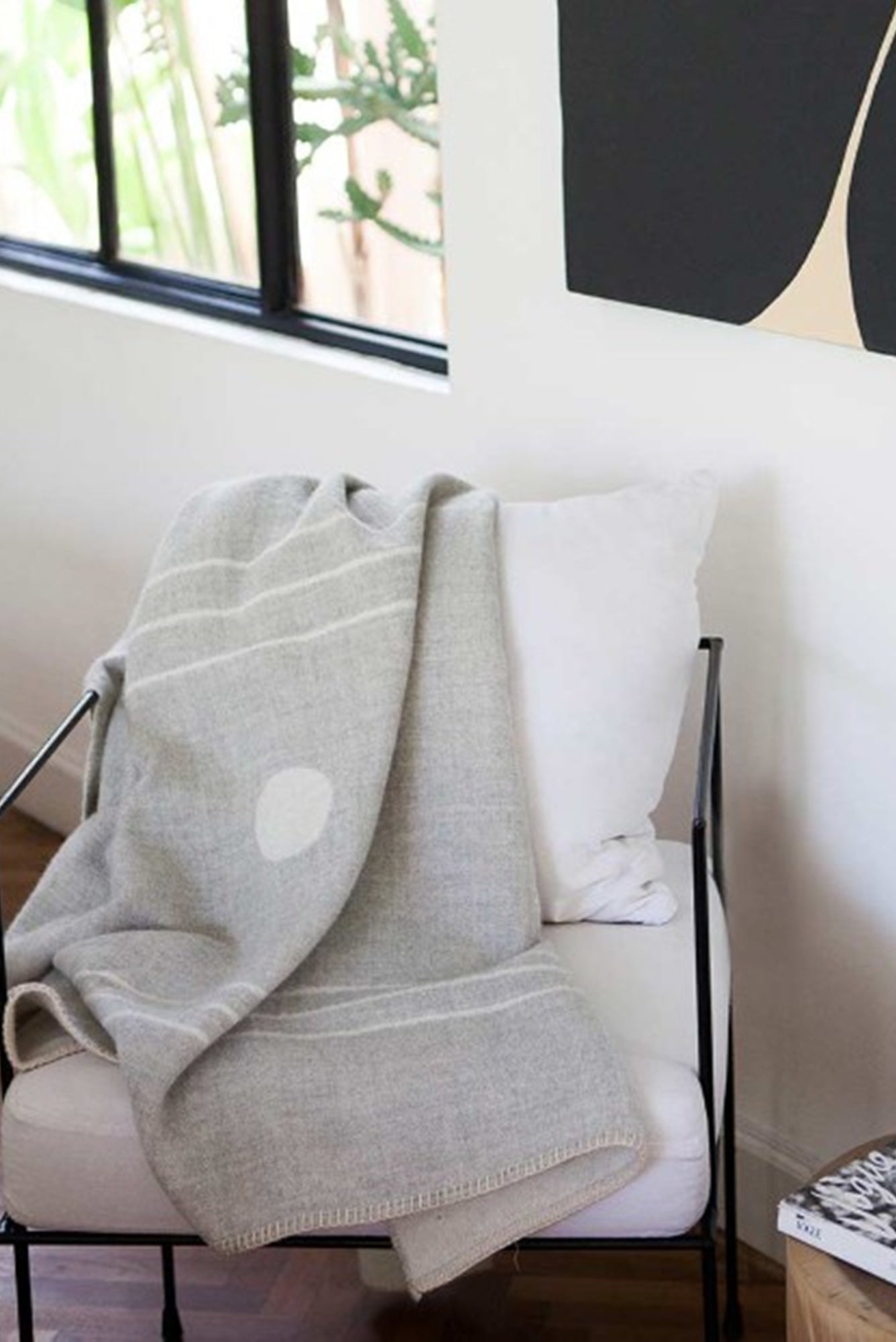 Gray blanket draped over a chair with a white pillow in a room with a window and abstract art.