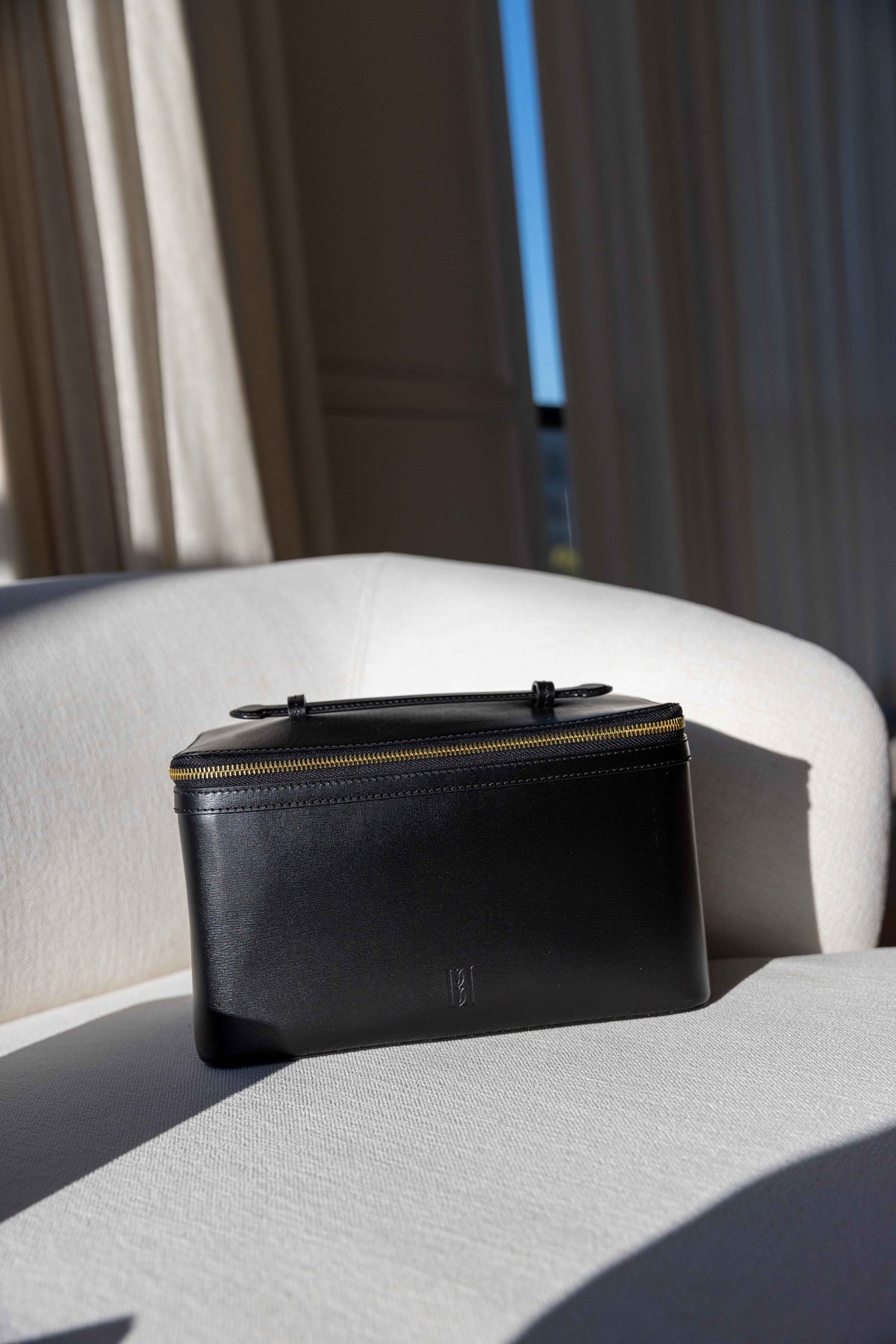by Malene Birger Aya Beauty Bag in black leather with gold zip and top handle, displayed on a white sofa