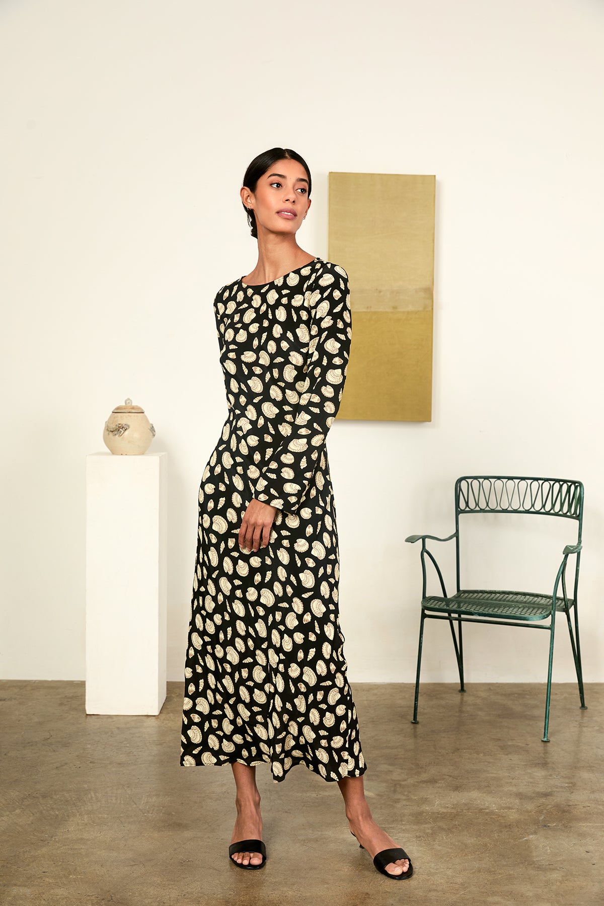Woman in a patterned dress standing in a room with a chair and wall art.