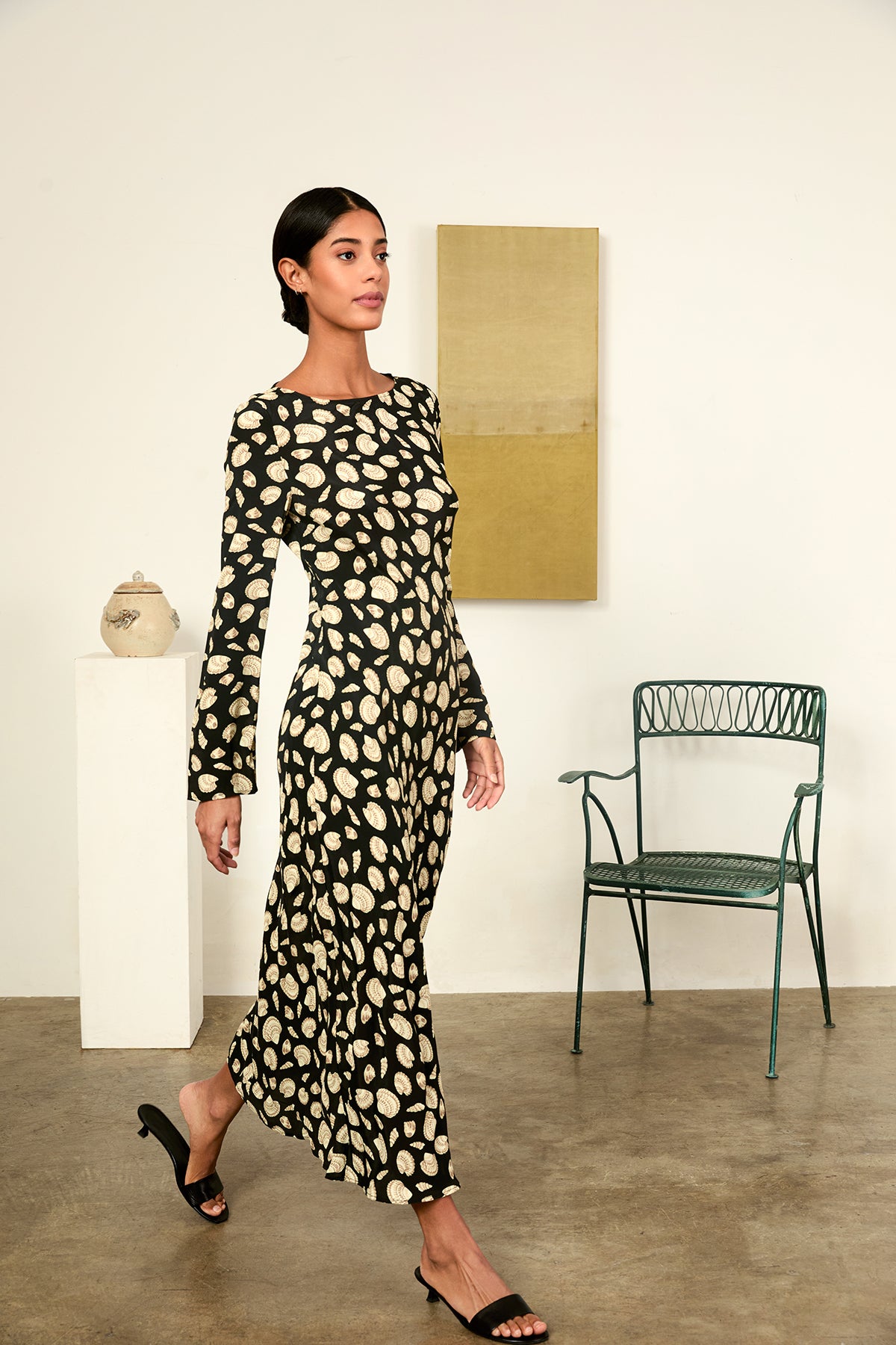 Woman in a patterned dress walking in an indoor setting with a chair and artwork in the background.