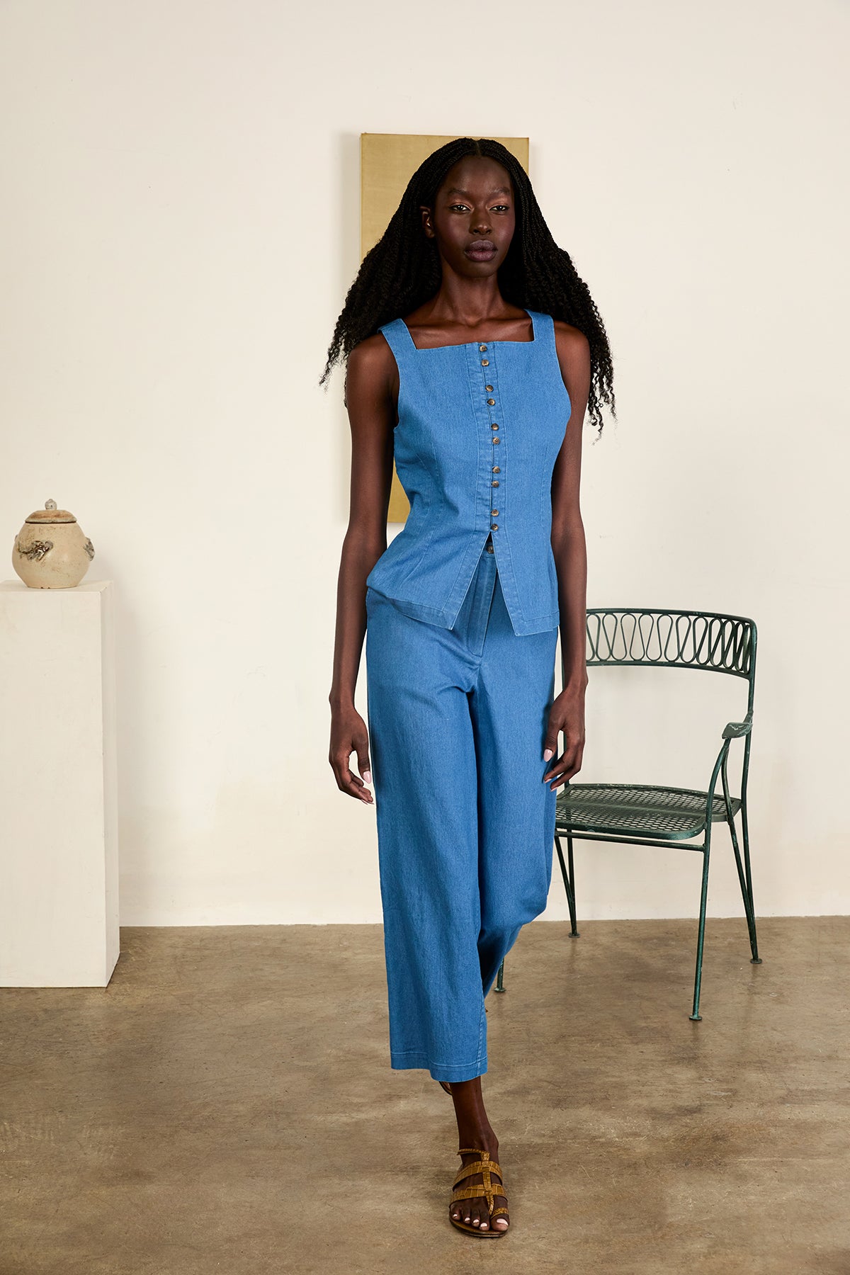 Woman wearing a blue outfit standing in a minimalistic room with a chair and decorative items.