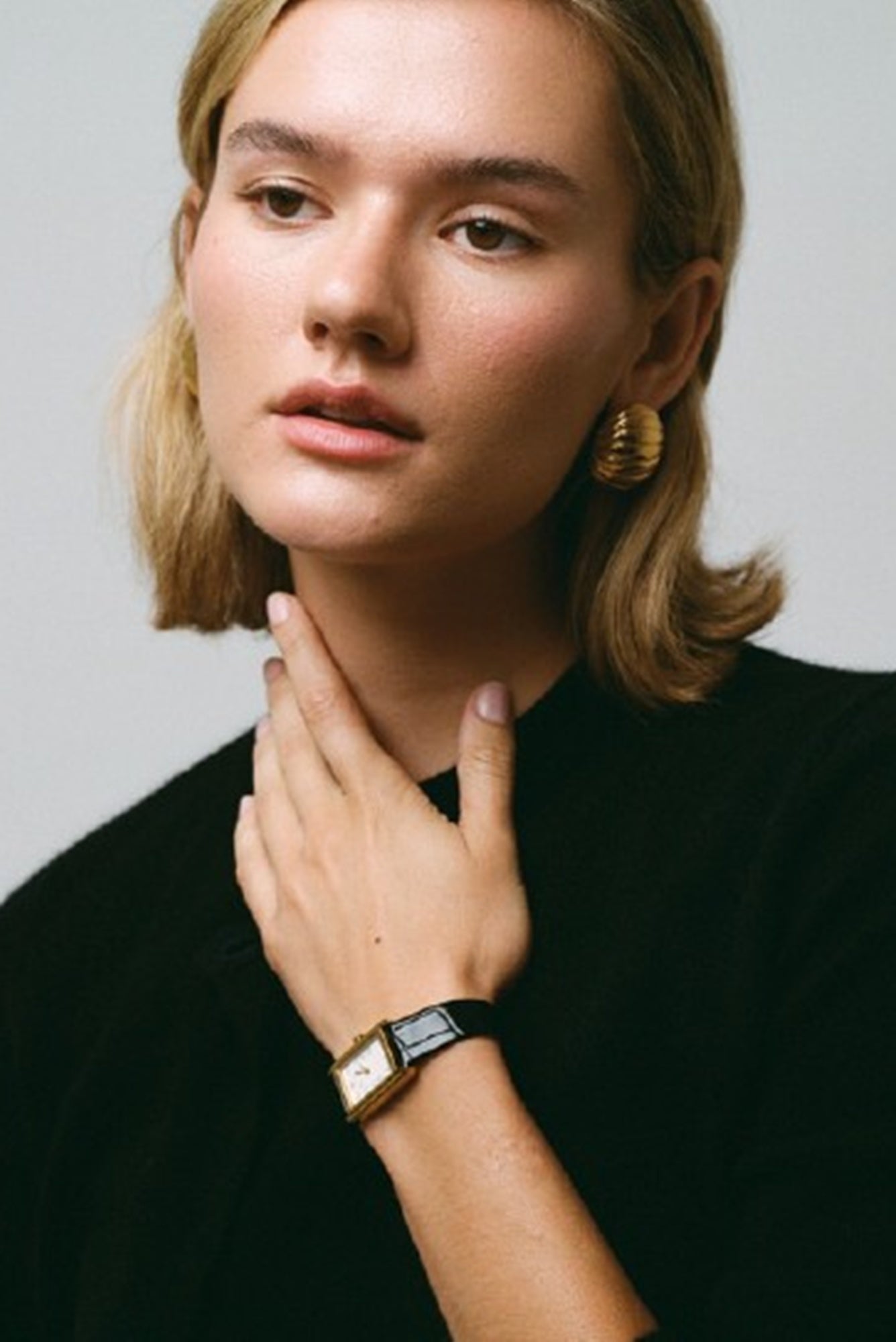 Woman wearing gold earrings and a watch on a plain background