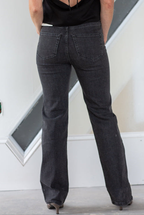 Person wearing a black open-back top and dark jeans standing against a white wall.